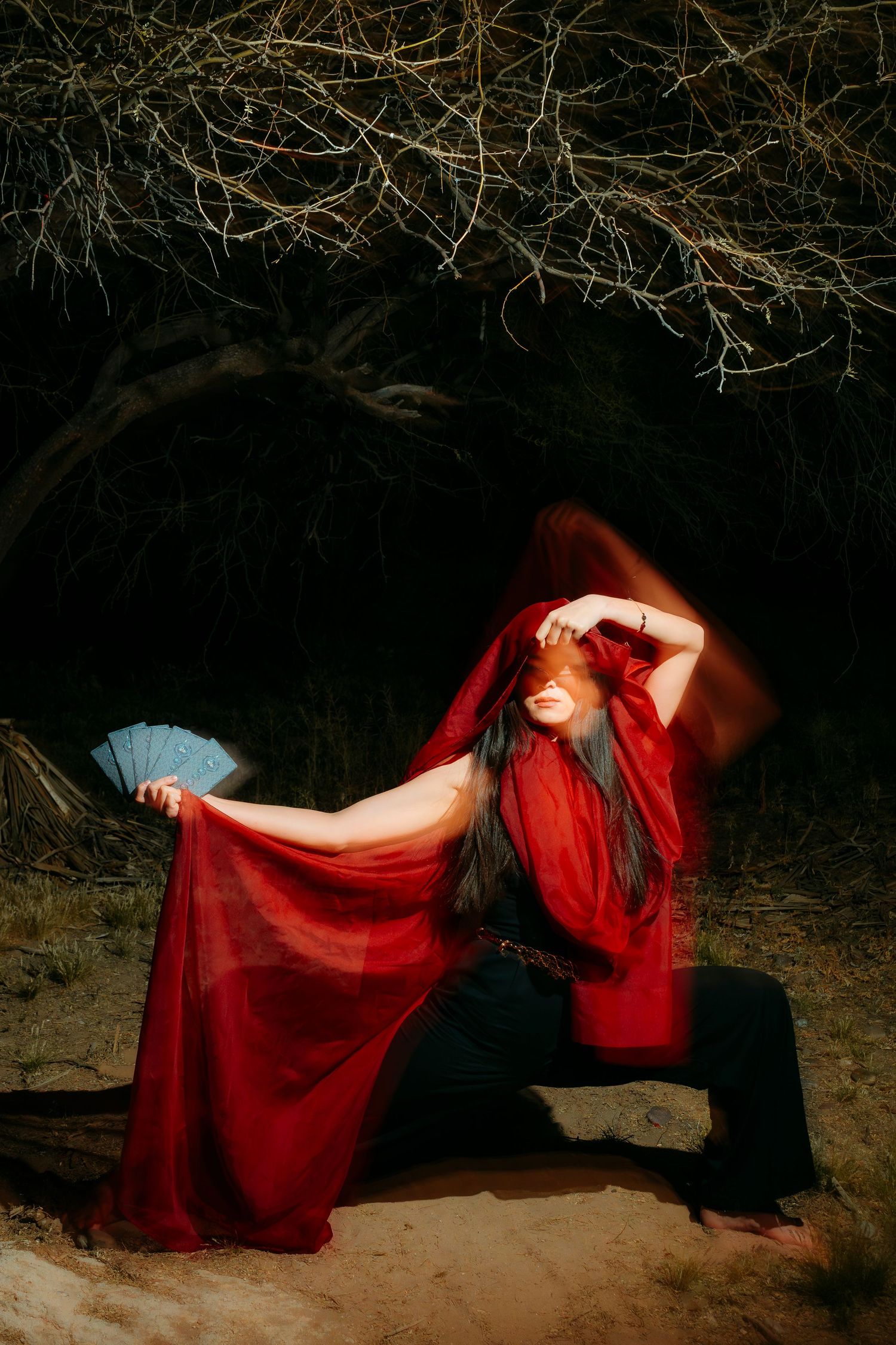 Woman in red hooded cape holds blue fan, seated outdoors at night near bare trees in dramatic pose.