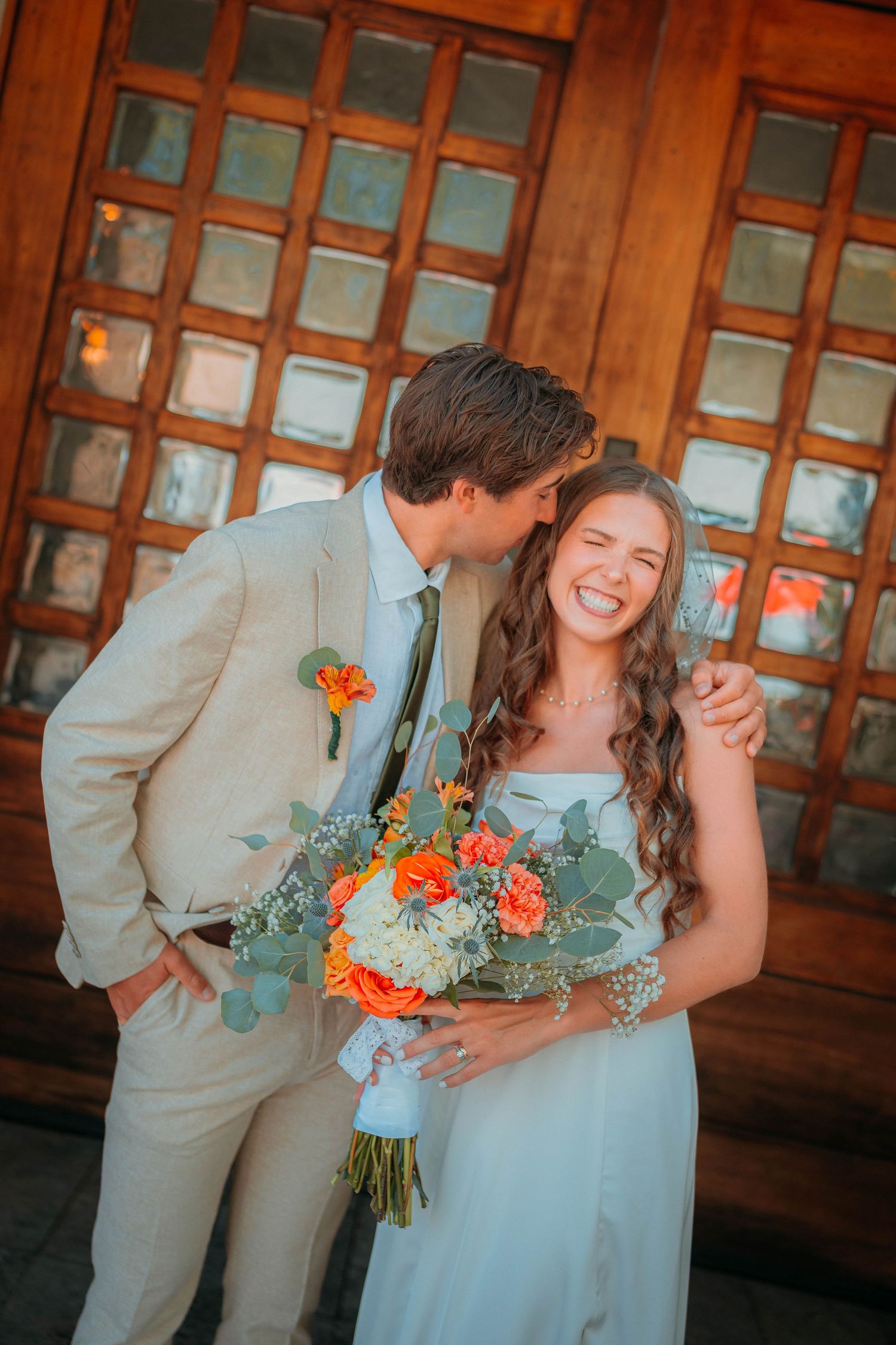 Smiling bride holding orange bouquet as groom kisses her cheek before wooden doors at Adero Scottsdale.