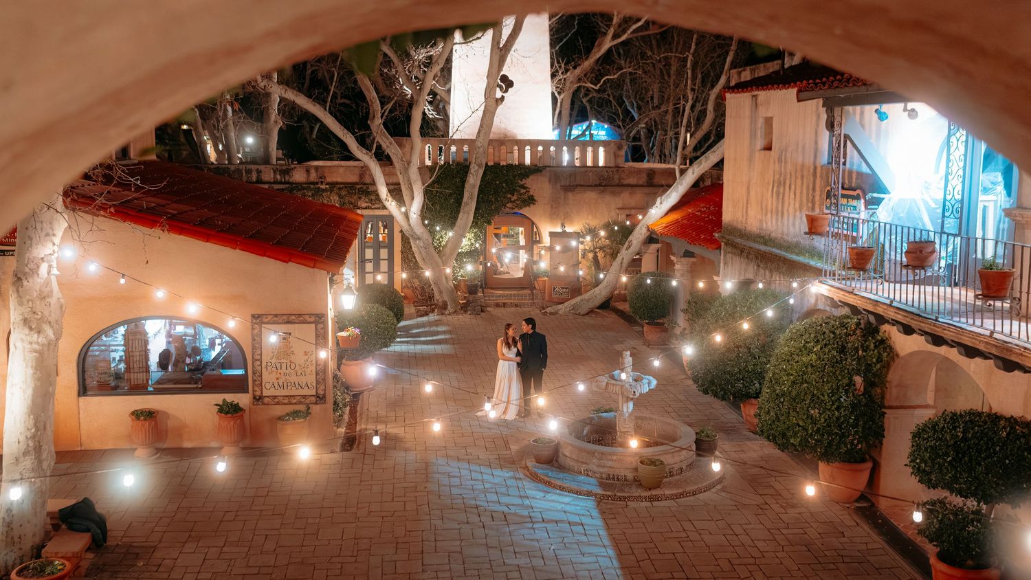 Couple shares romantic moment in beautifully lit courtyard with string lights and arched architecture at night.