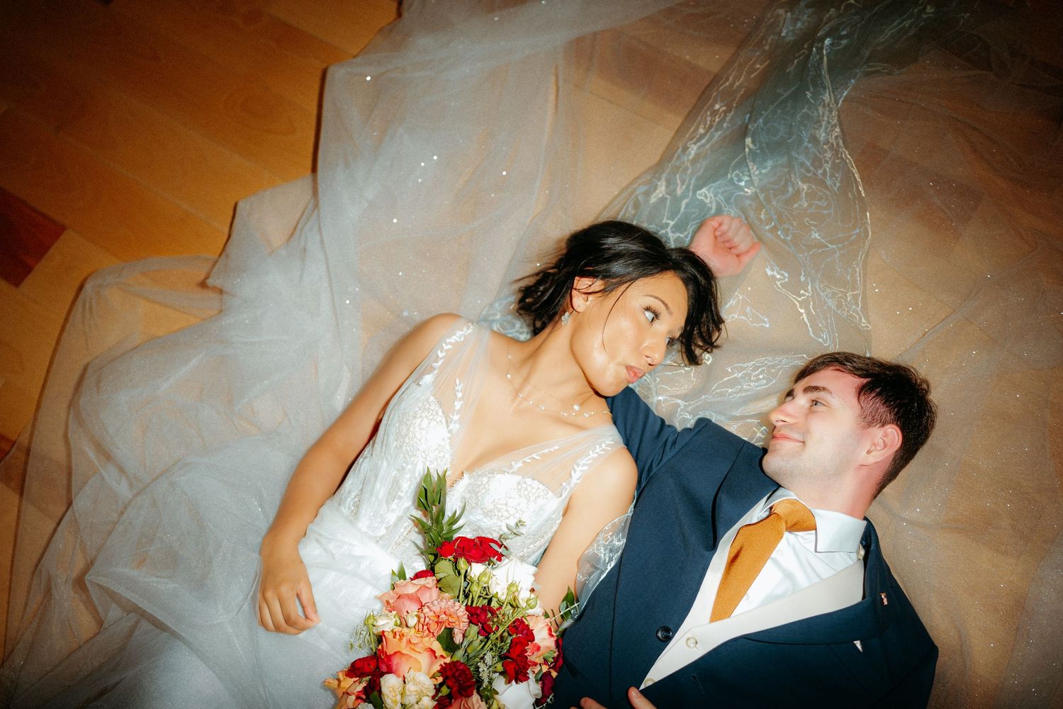 Bride and groom lying on floor under wedding veil, smiling at each other, bride holding colorful bouquet.