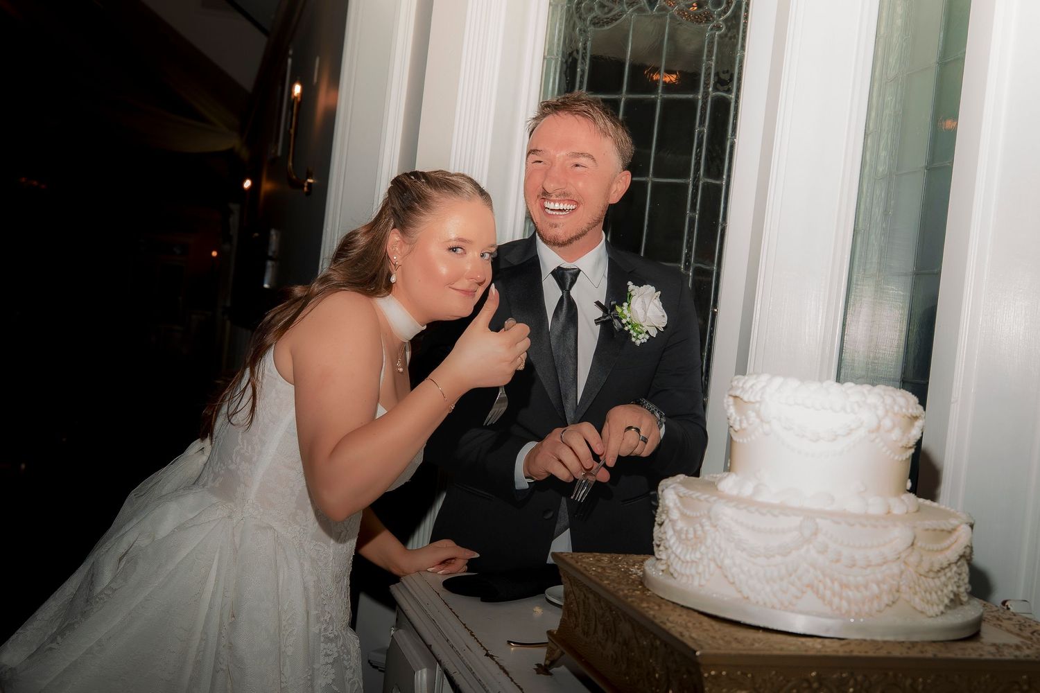 Bride and groom laughing while cutting their white wedding cake at a nighttime reception.