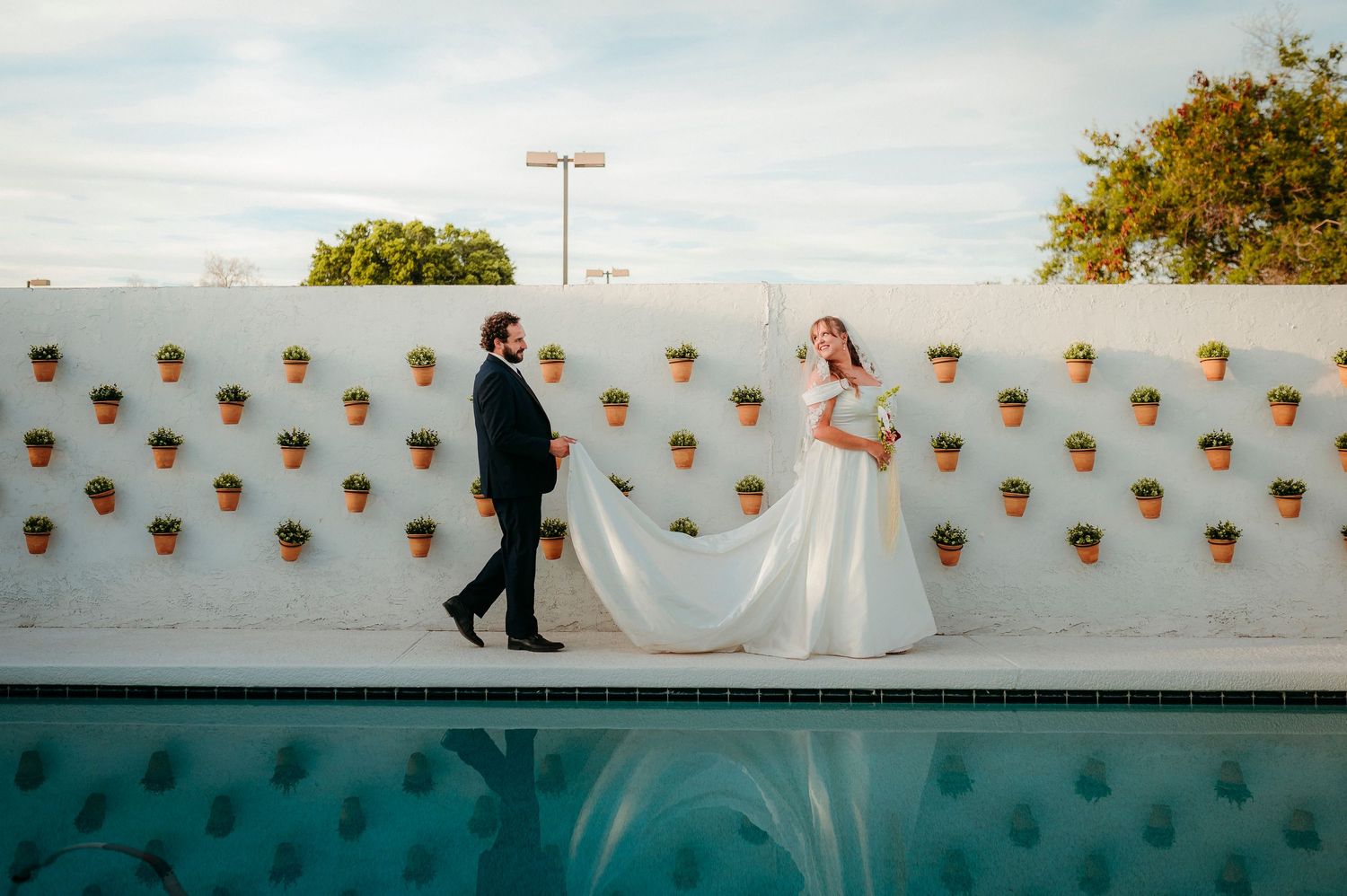 Bride and groom walk along poolside white wall decorated with terracotta pots at wedding venue.
