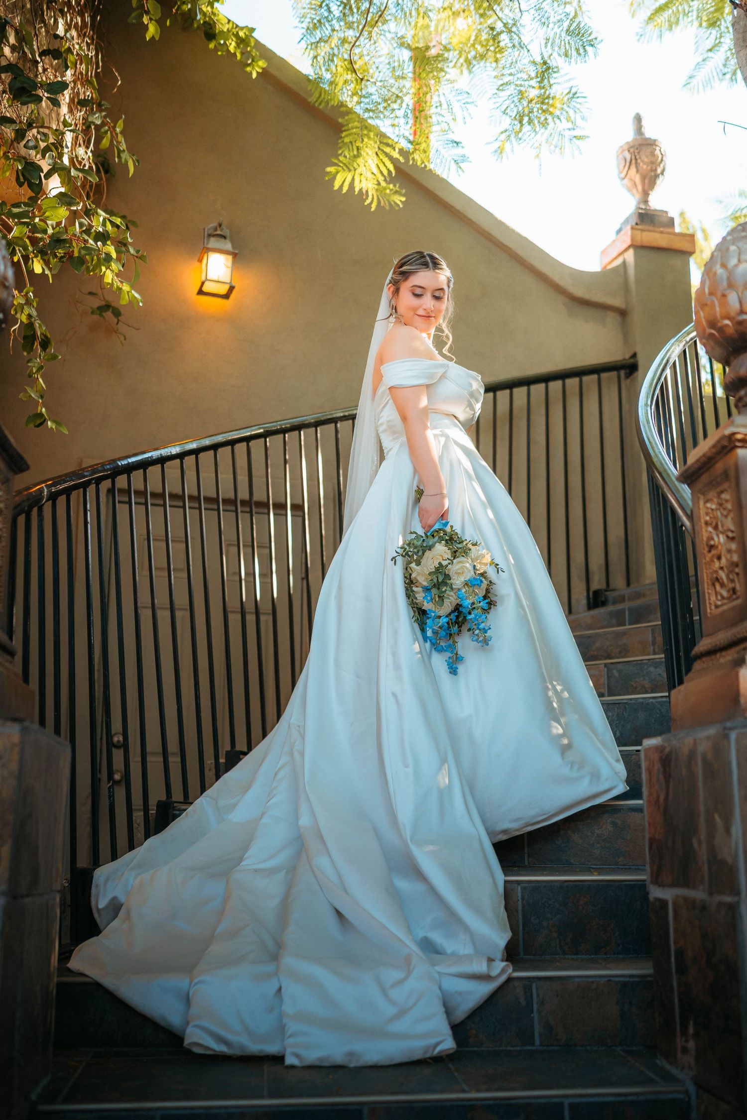 Elegant bride in off-shoulder white ball gown with blue bouquet posing on outdoor staircase at golden hour.
