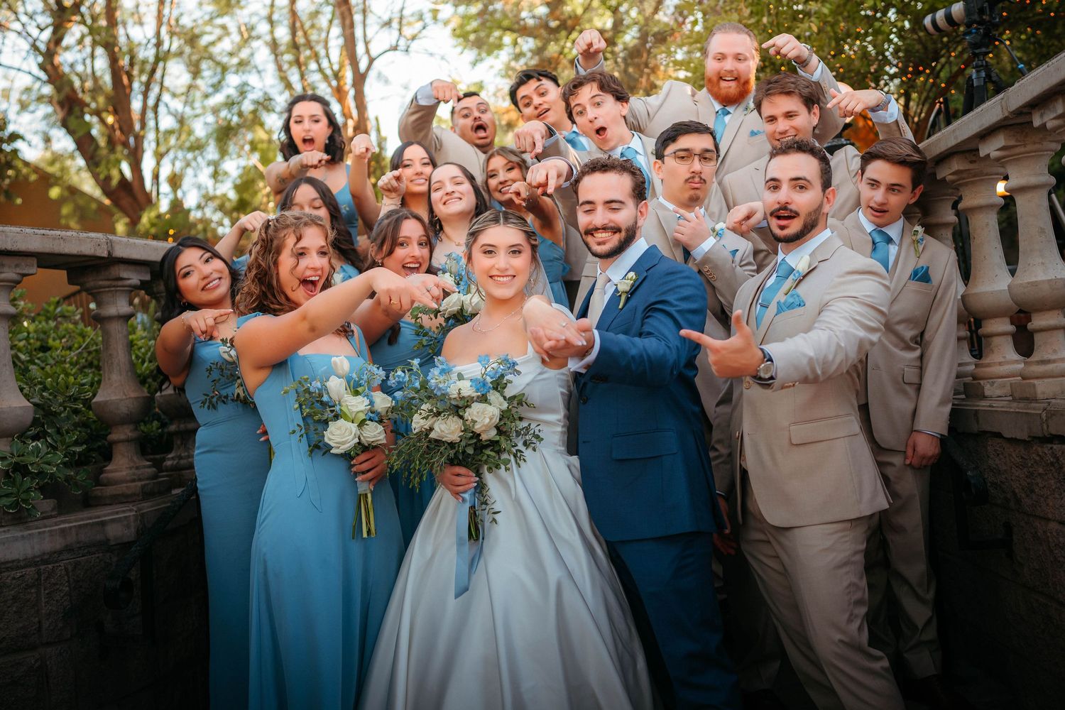 Joyful wedding party in blue bridesmaid dresses and tan suits celebrating with the bride and groom outdoors.