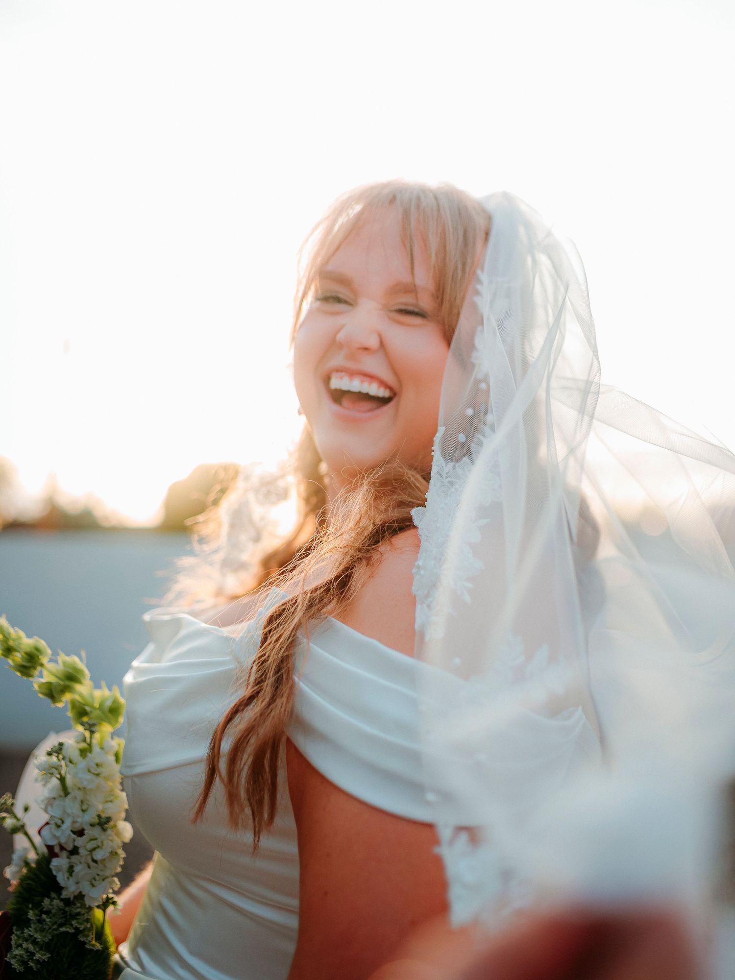 Joyful bride laughing in golden sunlight wearing a white wedding dress and veil, holding a bouquet of white flowers.