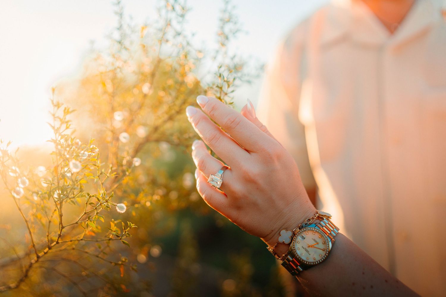 Close-up of a woman's hand wearing a diamond engagement ring and gold watch, surrounded by golden sunlit foliage.
