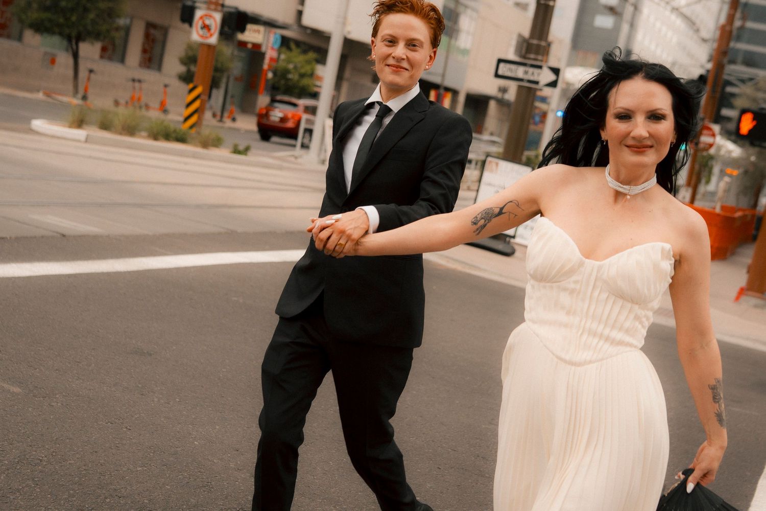 A bride in a white gown and groom in a black suit hold hands while crossing a city street on their wedding day.