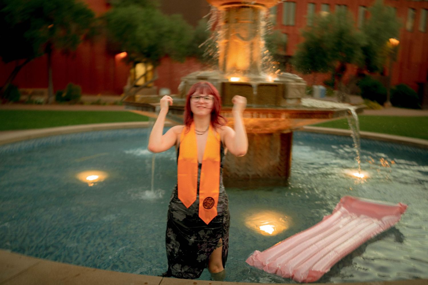 Graduate in orange sash celebrates in fountain at dusk near red brick building with inflatable float nearby.