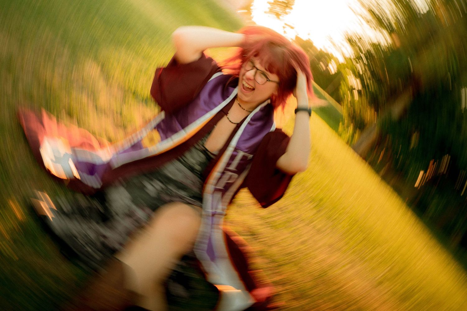 A joyful woman with red hair and glasses spins outdoors in golden sunlight, wearing a purple jacket.
