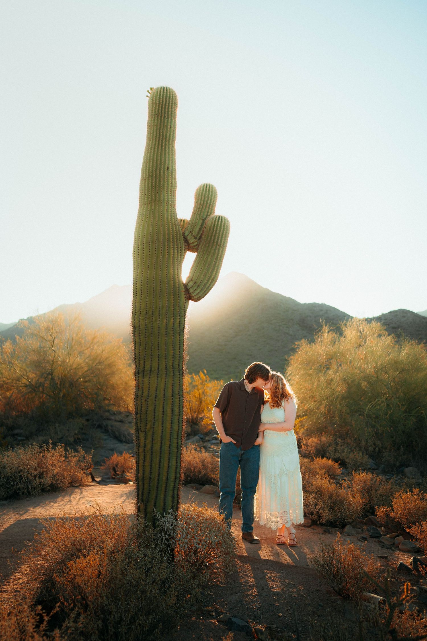 Couple sharing a romantic moment beside a tall saguaro cactus in the Arizona desert at sunset.