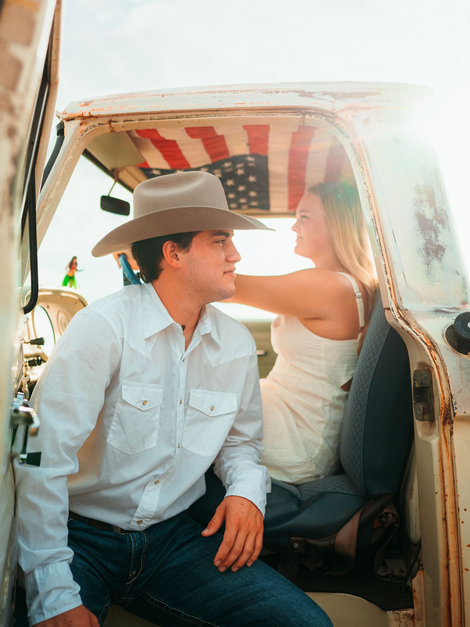 A couple sits in a vintage truck with an American flag backdrop, the man in a cowboy hat gazing at a woman in a white dress.