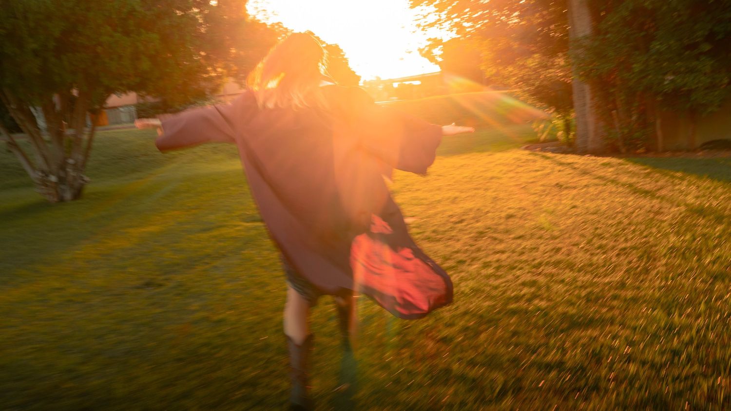 Child in a cape runs through a sunlit green yard at golden hour, backlit by warm glowing sunset light.