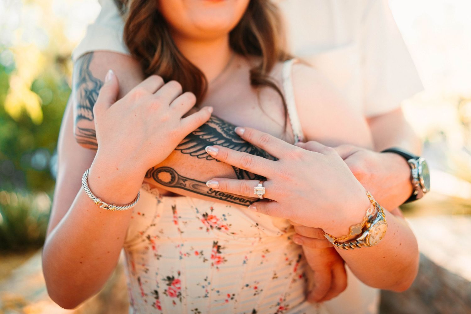 Couple embracing outdoors, woman in floral dress with tattoos and bracelets, man in white shirt wearing a watch.