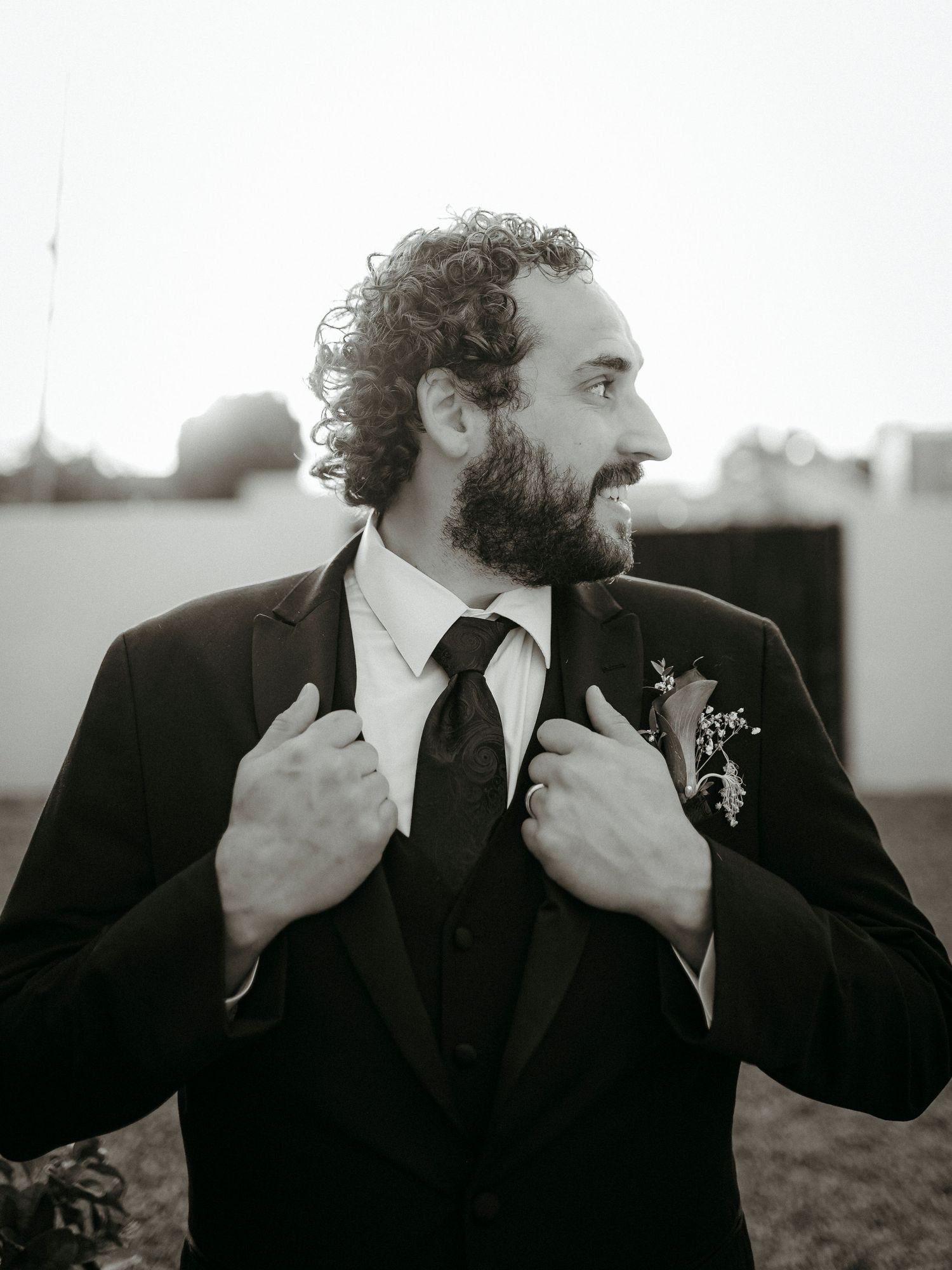 Black and white portrait of a bearded groom in a suit adjusting his lapels with a boutonniere, smiling outdoors.