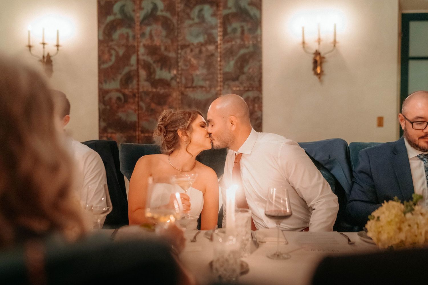 Newlywed couple sharing a romantic kiss at their wedding reception dinner table with candlelight ambiance.