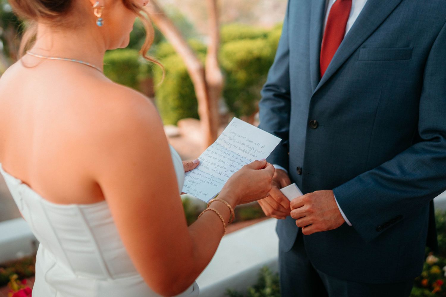 Bride reads handwritten wedding vows while holding grooms hand during outdoor ceremony.