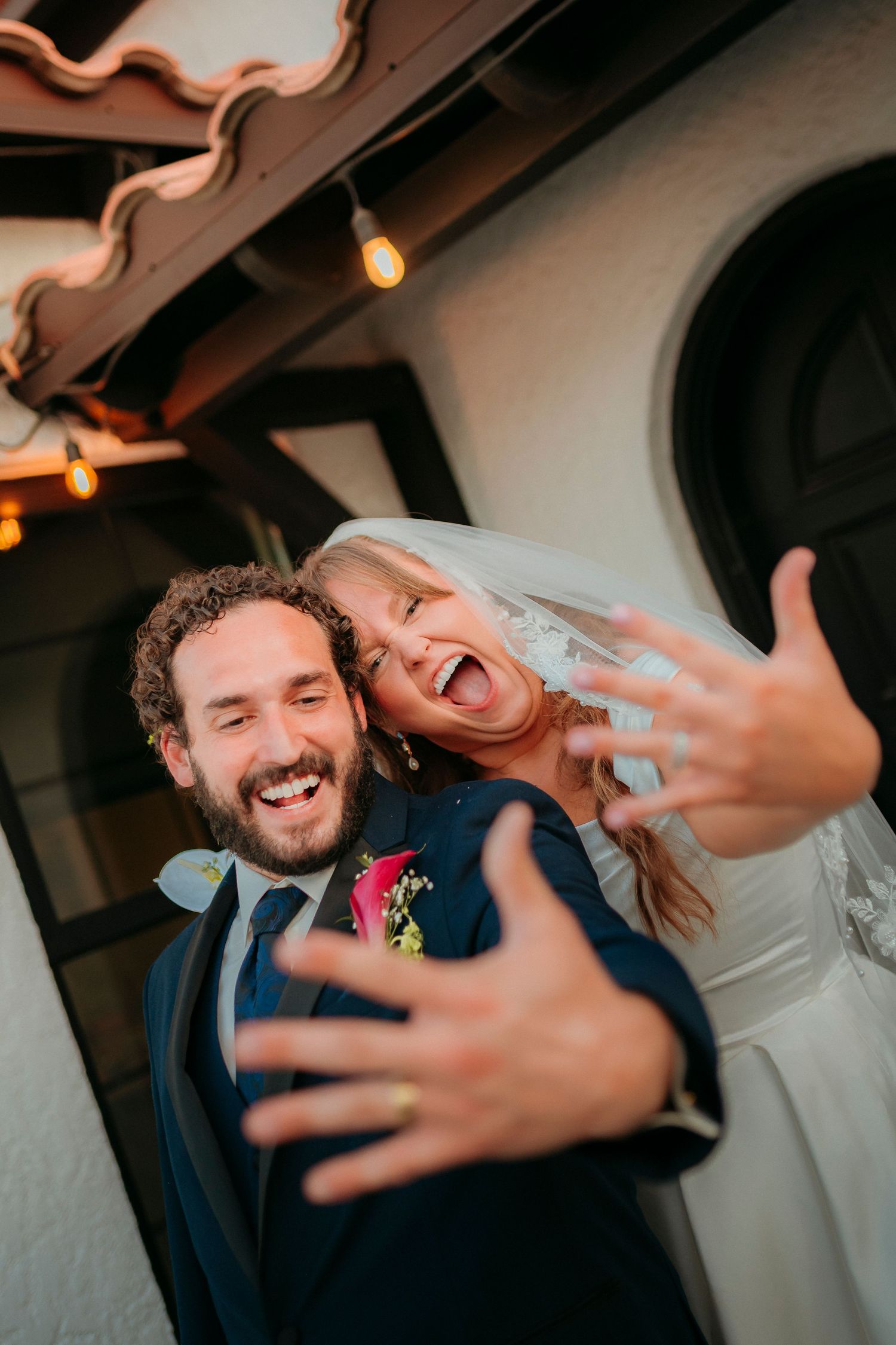 Excited newlyweds laugh and show off their wedding rings outside their reception venue with string lights above.
