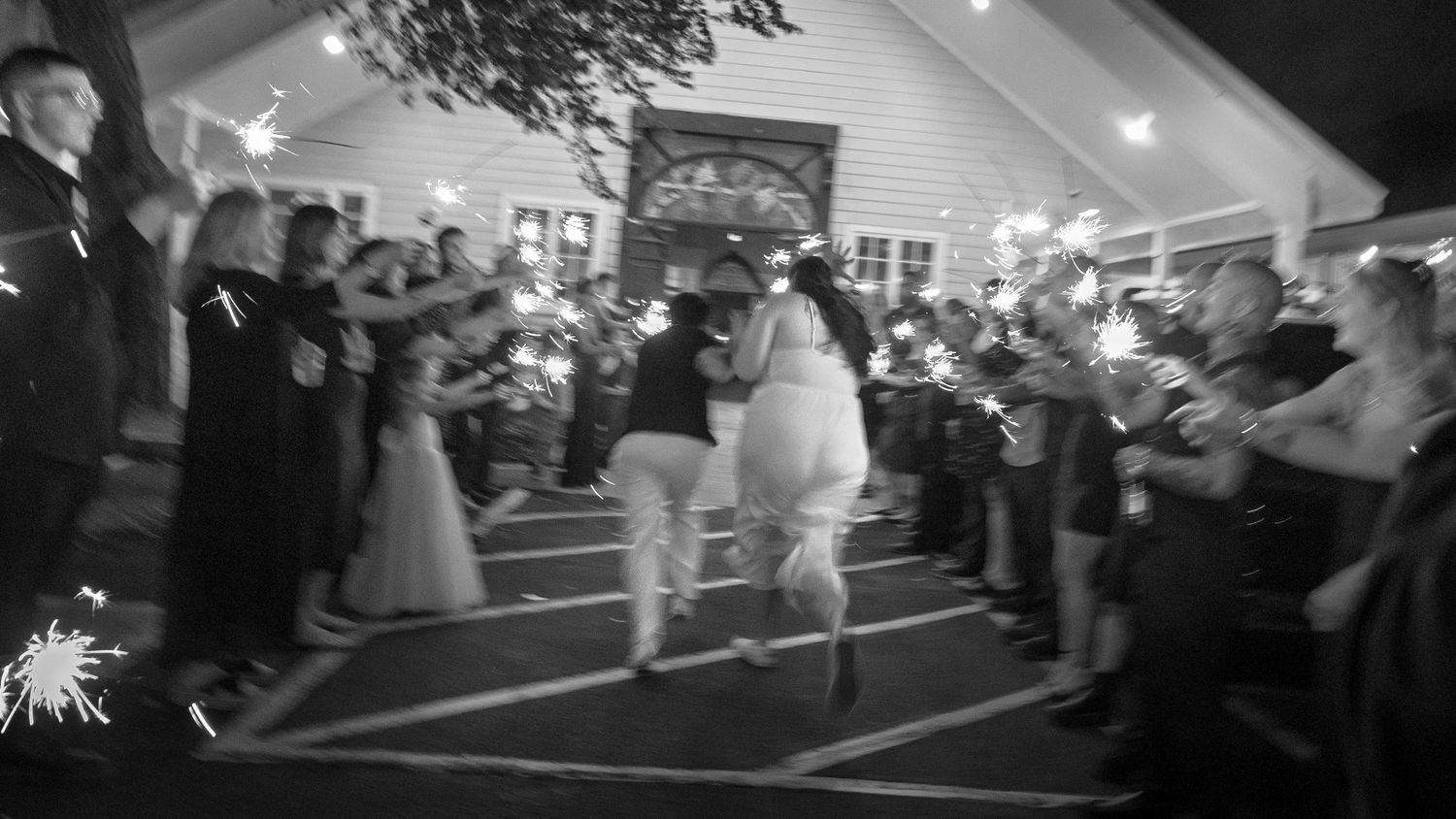 Black and white photo of newlyweds running through a sparkler send-off at their outdoor wedding reception.