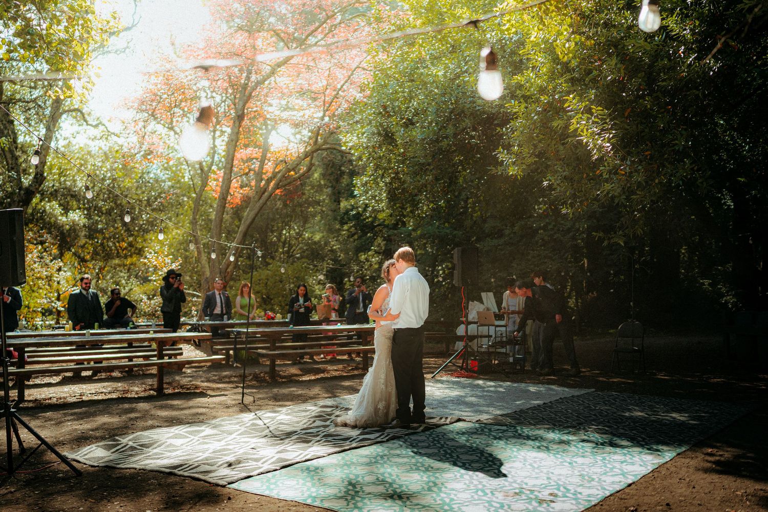 Couple shares first dance outdoors on patterned floor under string lights surrounded by lush trees at wedding reception.