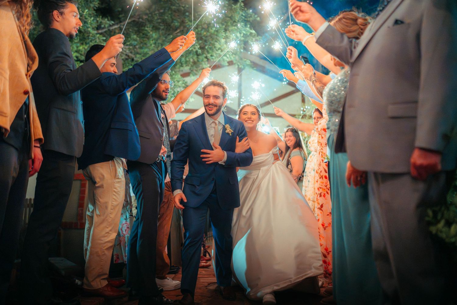 Bride and groom walking through a sparkler send-off tunnel surrounded by cheering guests at night.
