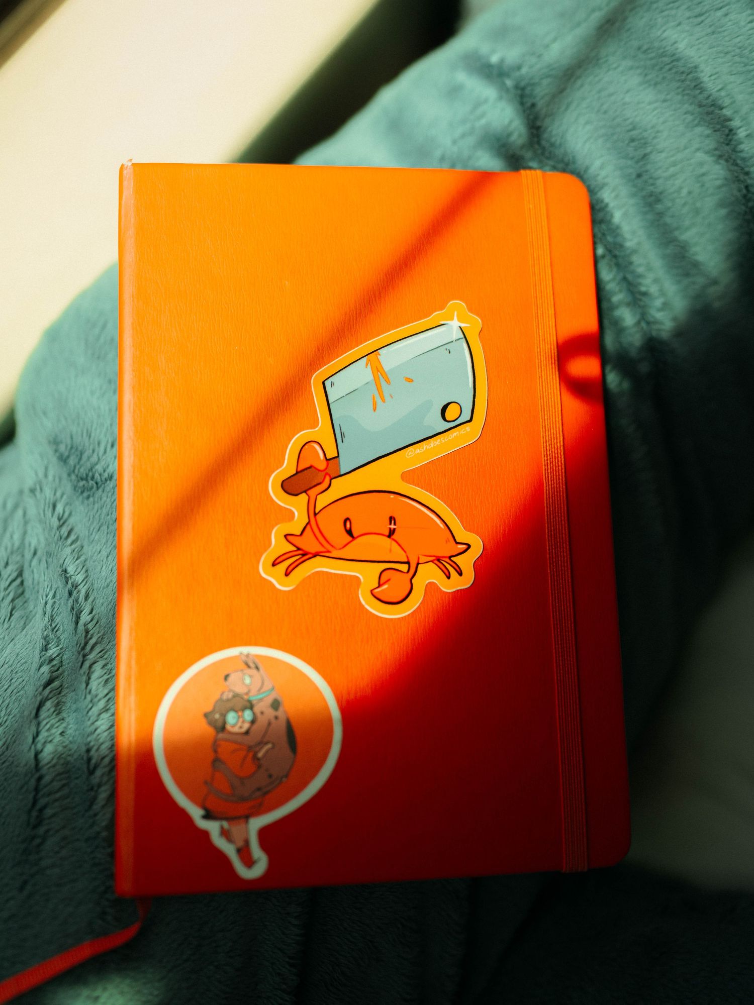 Orange Moleskine notebook with colorful cartoon Ash Does Comics crab sticker on teal fabric background.