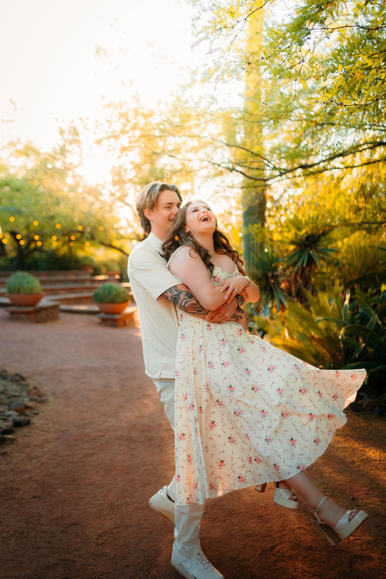 Couple dancing joyfully at golden hour in a lush garden, woman in floral dress laughing as man lifts her.