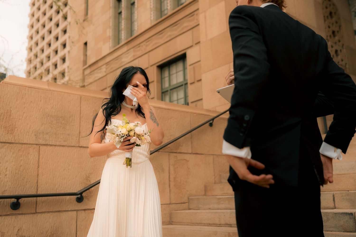 Emotional bride in white gown holds bouquet and wipes tears while groom in black suit stands before her on stone steps.