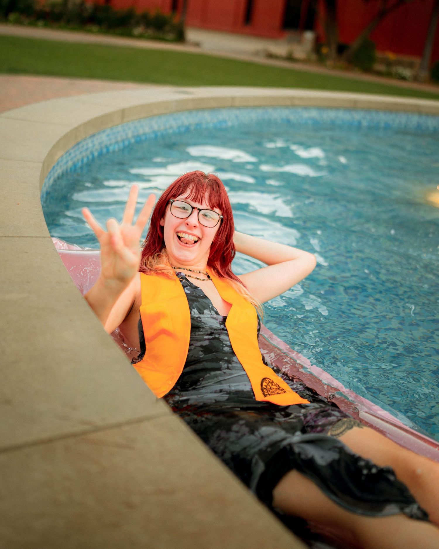 A cheerful woman with red hair and glasses in a black dress and orange vest sits by a pool, waving.