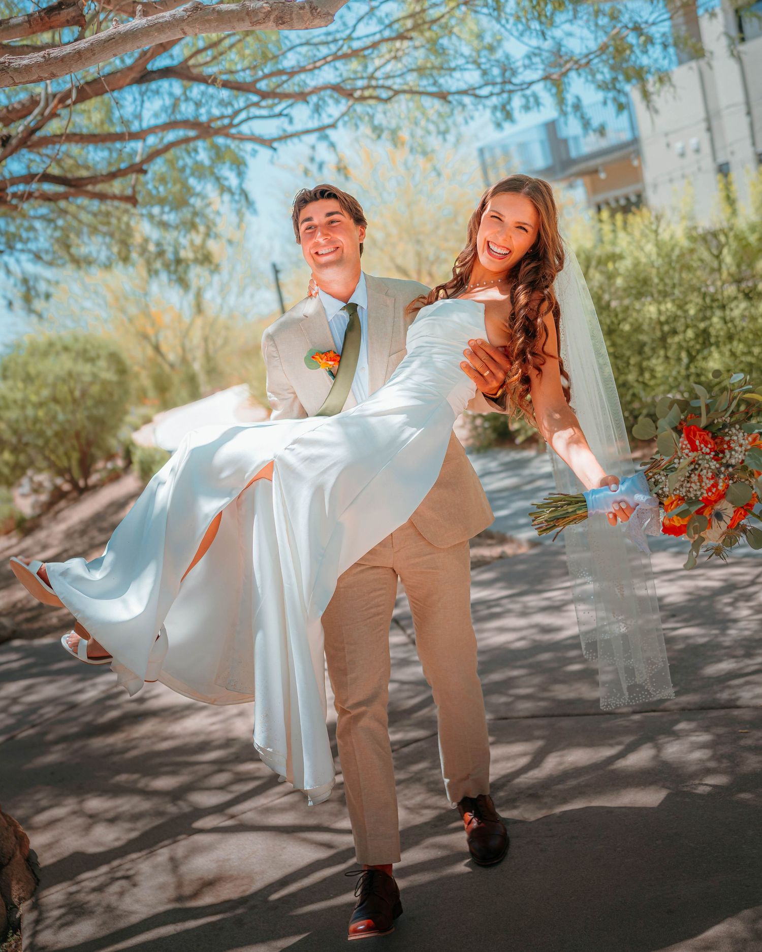 Joyful groom carries laughing bride in white wedding dress outdoors under trees at sunny desert wedding.