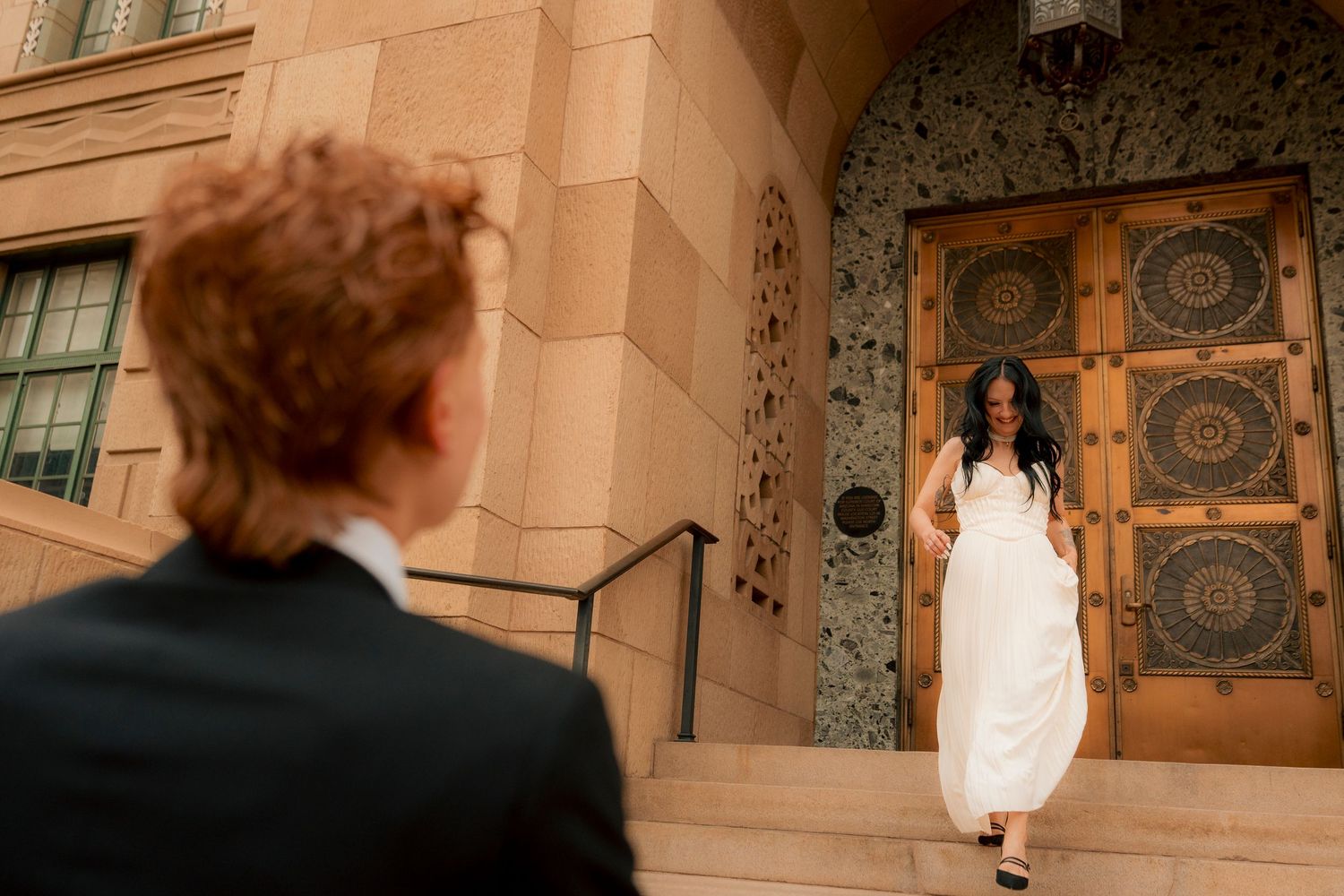 Bride in white dress stands at ornate building entrance as groom in black suit approaches on steps.
