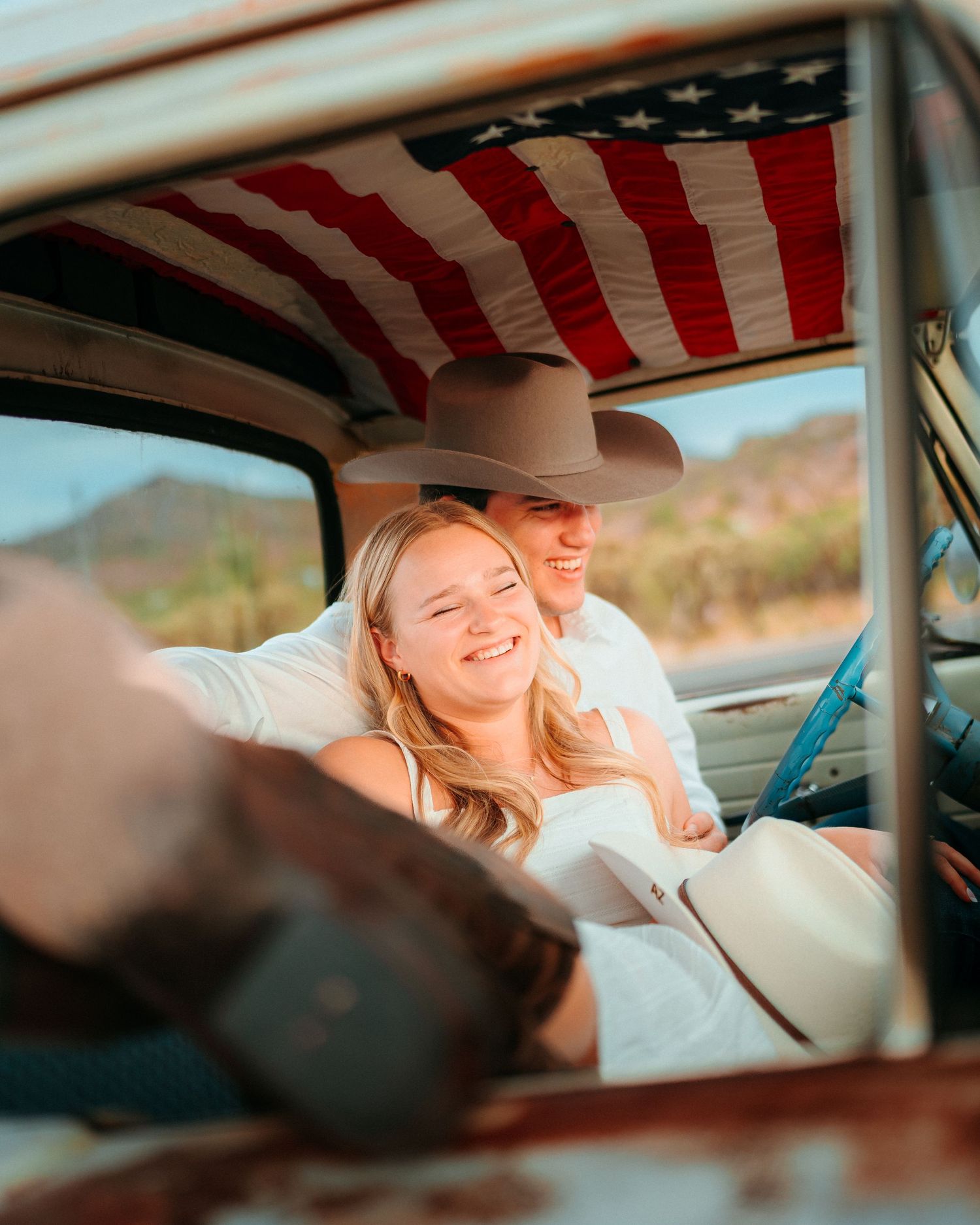 Couple smiling in vintage truck cab with American flag lining, woman in white dress, man in cowboy hat, desert backdrop.