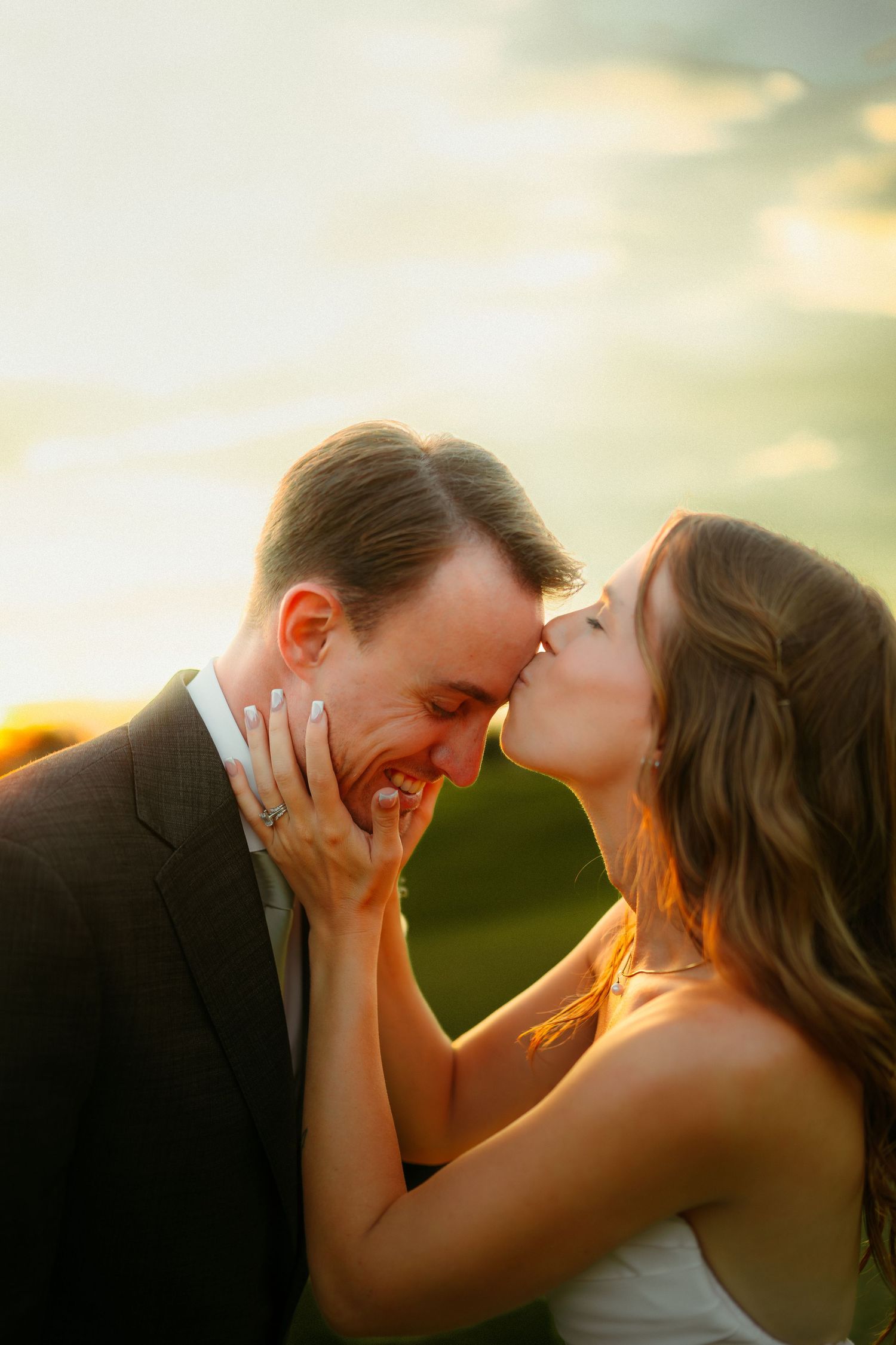 Bride kisses groom on cheek at golden hour, her diamond ring visible as they share a romantic outdoor wedding moment.