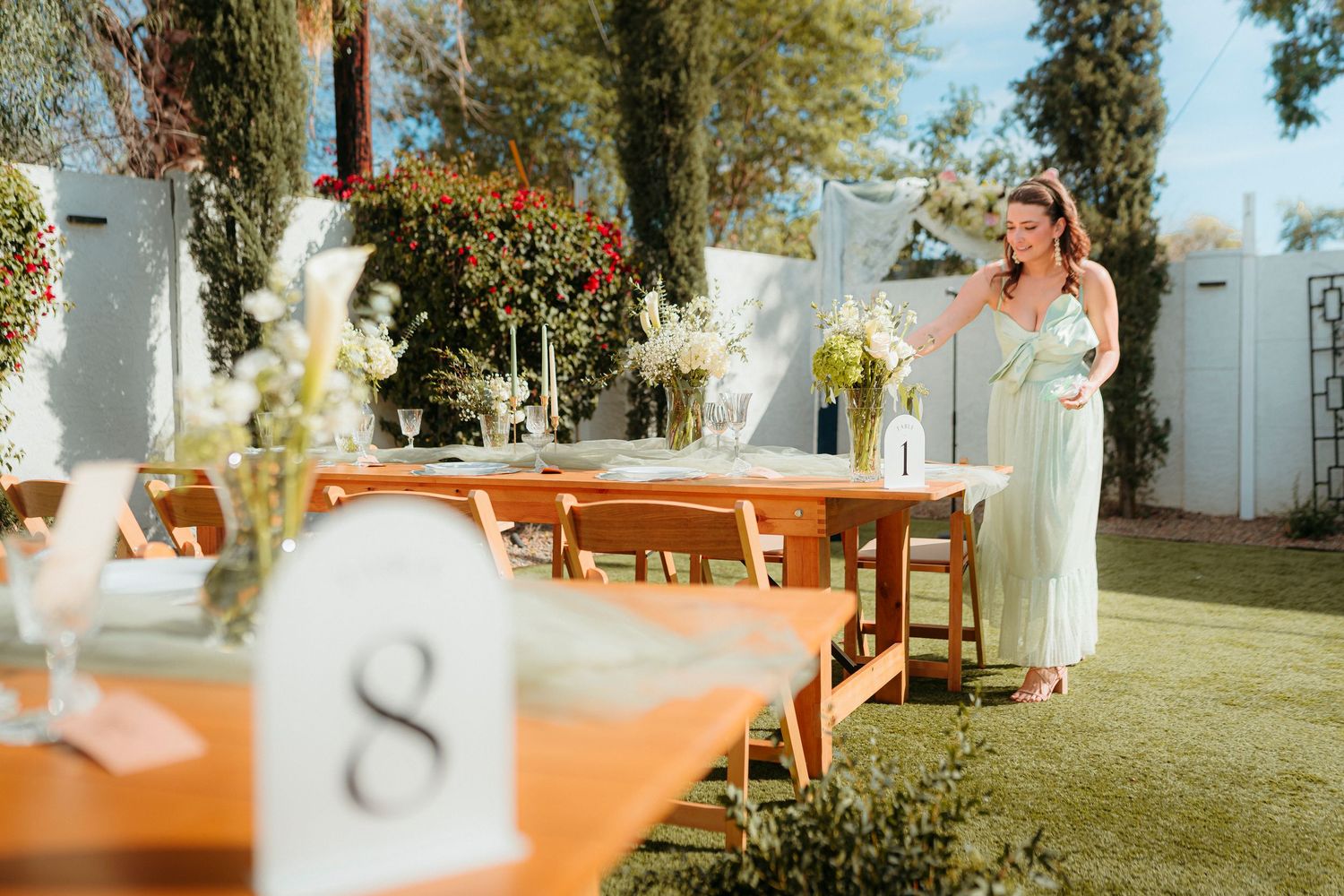 A woman in a green dress arranges floral centerpieces on wooden tables set up for an outdoor wedding reception.