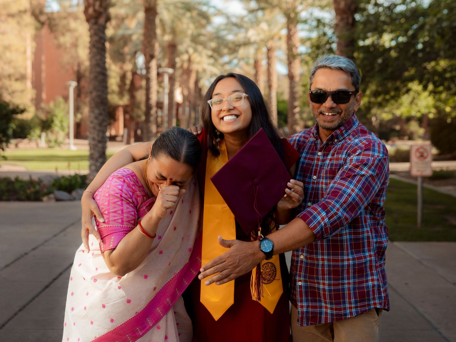 Proud ASU graduate in maroon gown embracing smiling parents on campus.