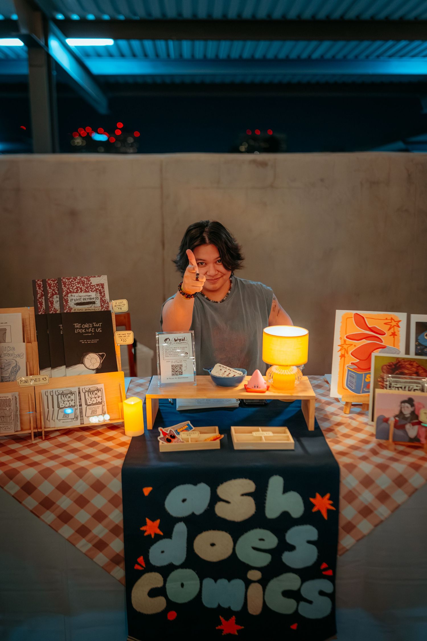 Artist Ash poses at their comic book vendor table with colorful artwork and a yellow lamp at a night market.