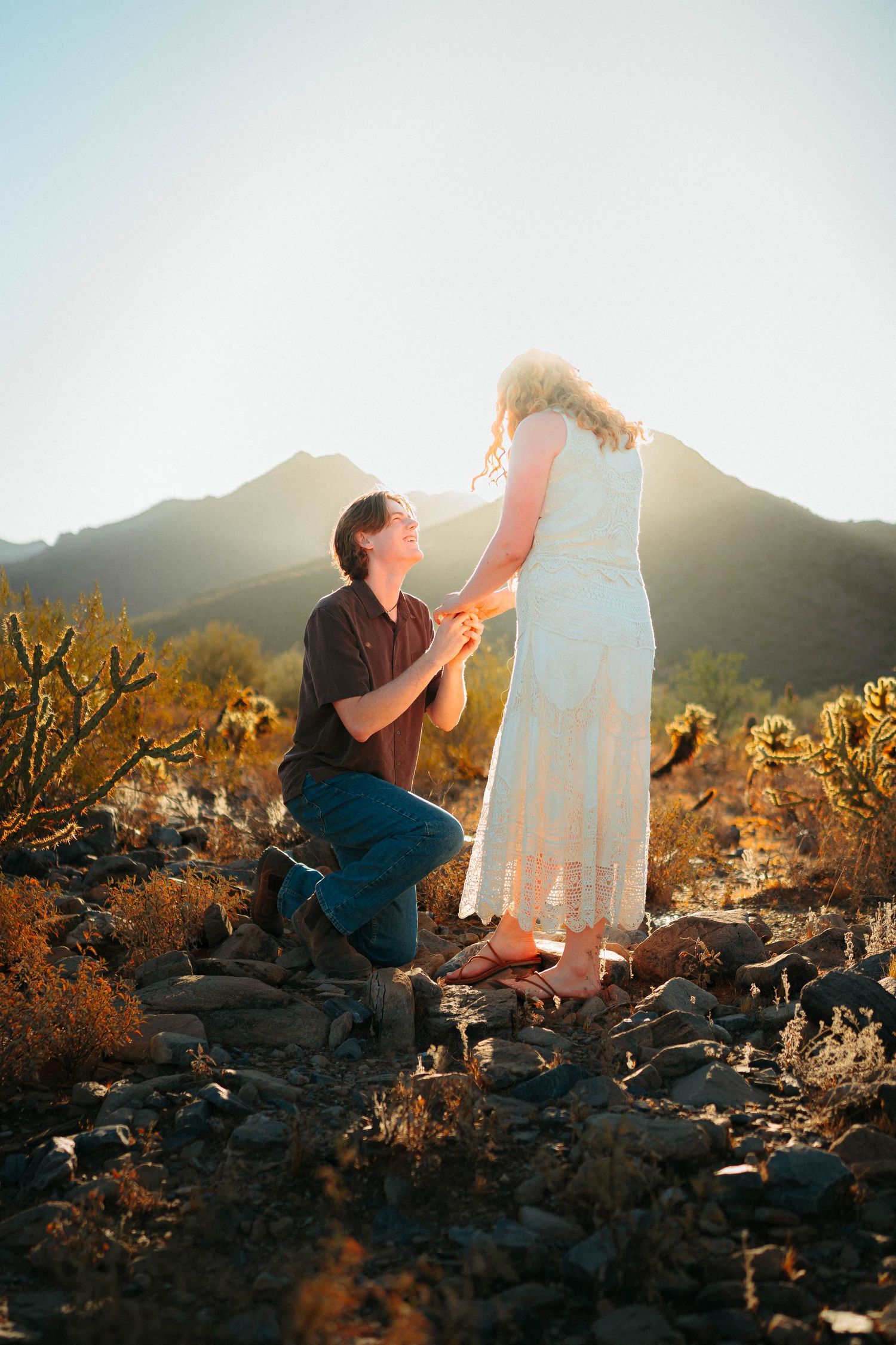 Man proposing on one knee to woman in white dress in rocky desert landscape at golden hour sunset.