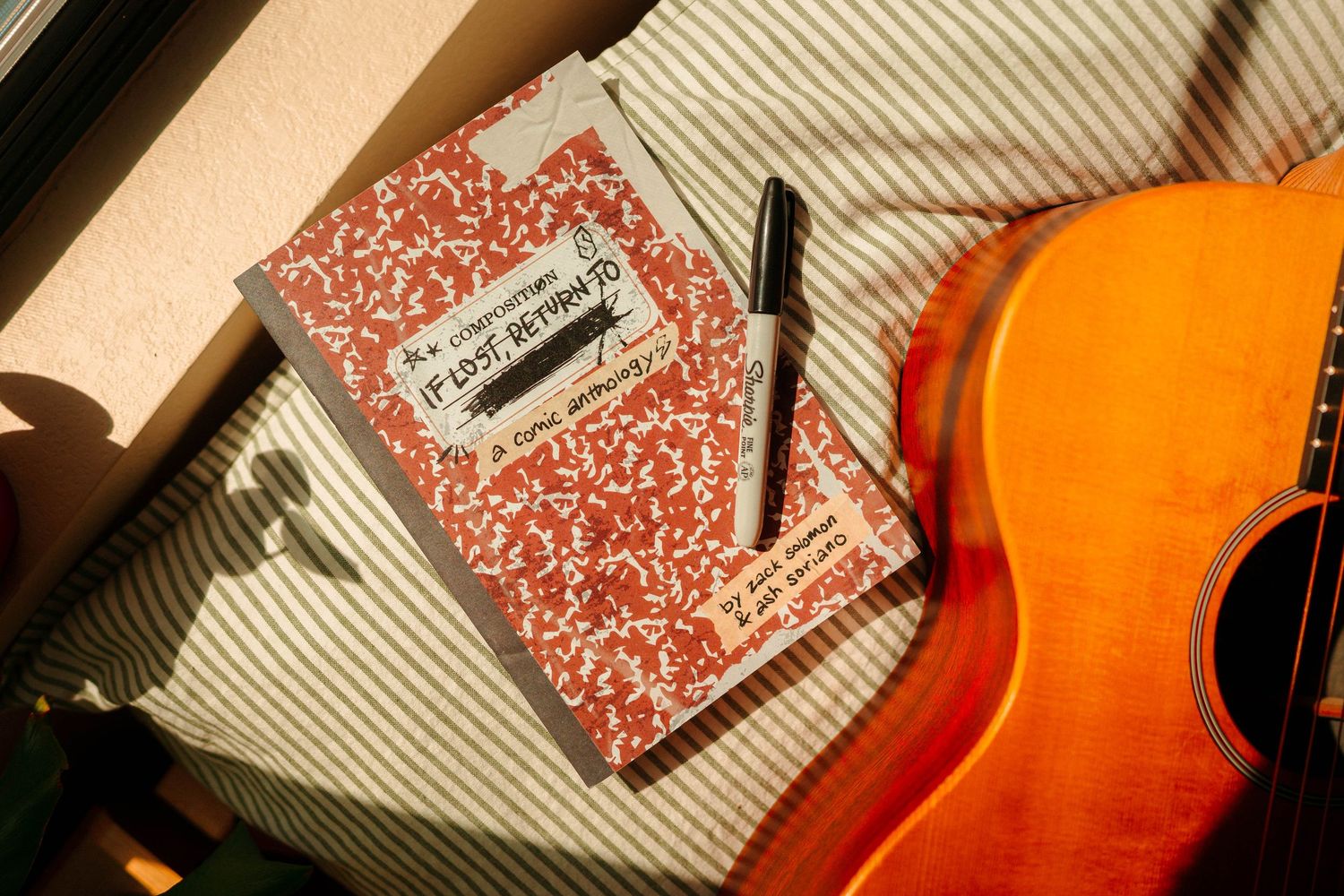 A red composition notebook with a pen rests beside an acoustic guitar in warm sunlight.