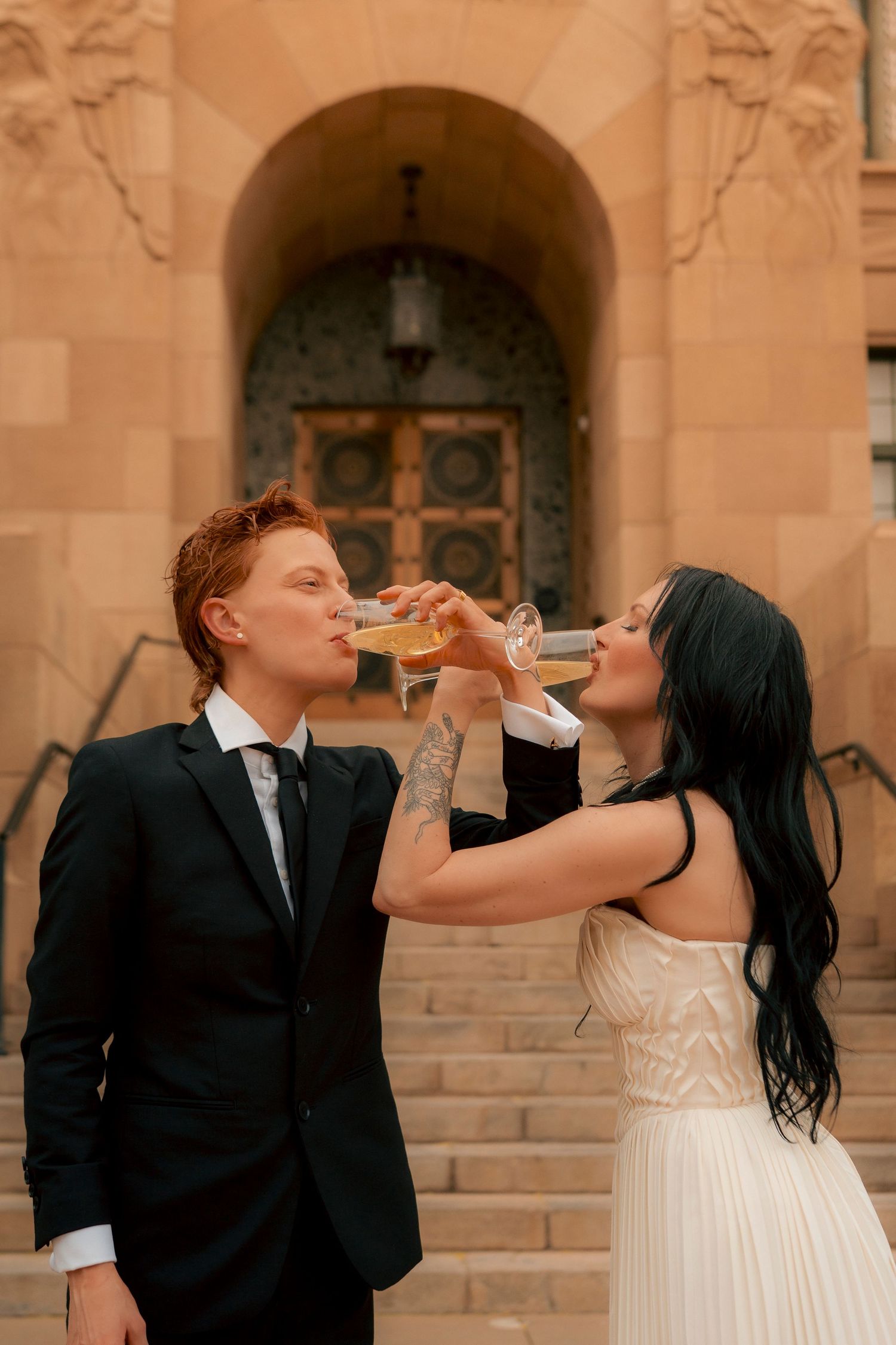Two newlyweds toast with champagne glasses on church steps, bride in white gown and groom in black suit.