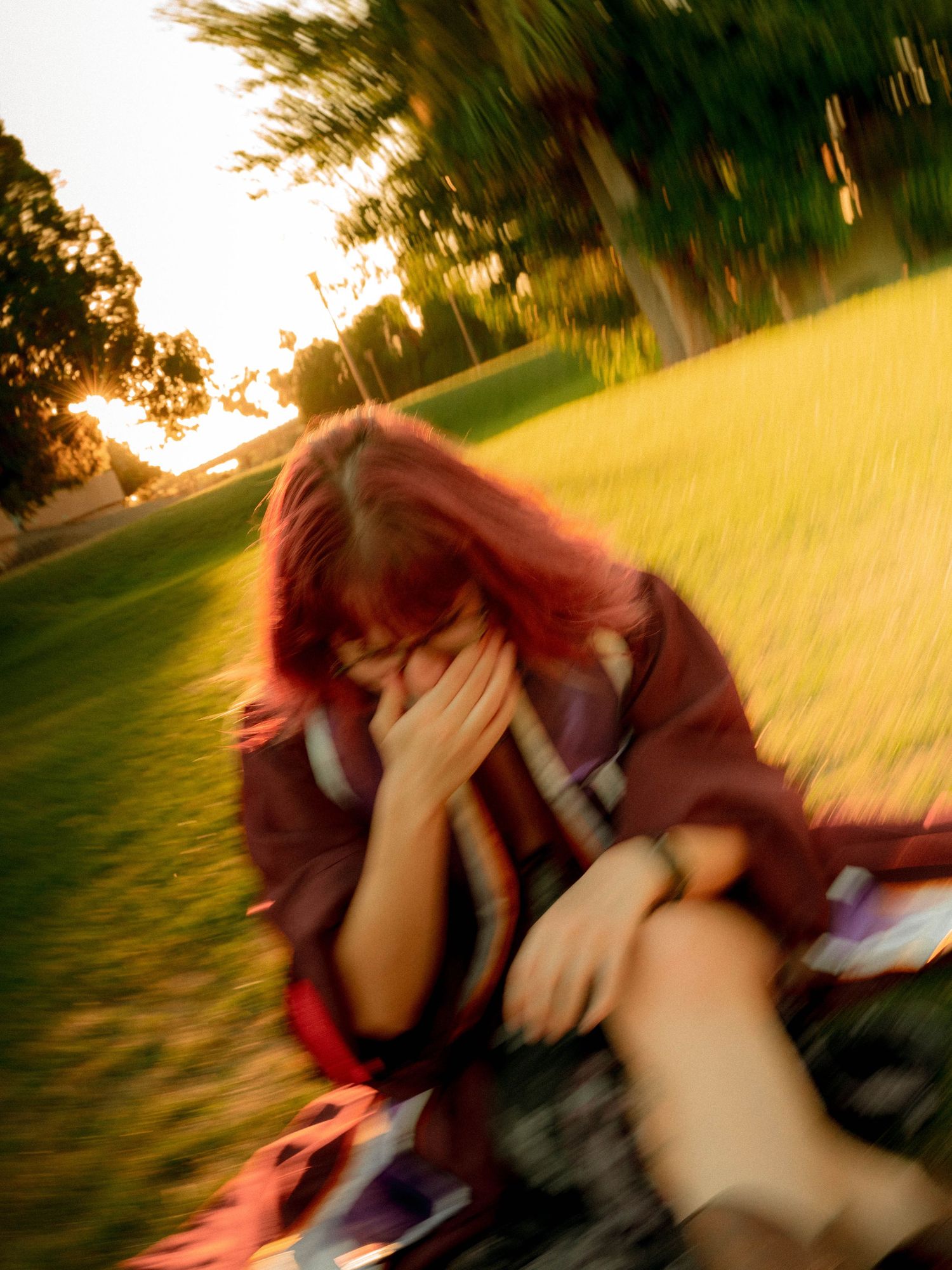 Red-haired woman sitting on grass in a park at sunset, covering her face with her hands in a blurred photo.