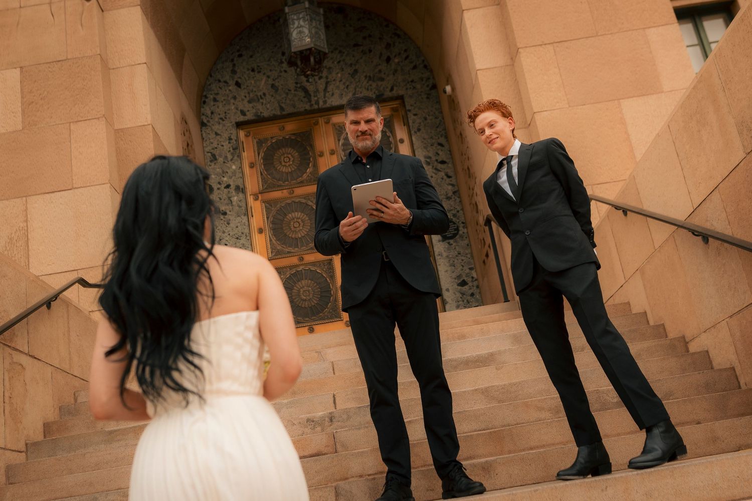Bride in white dress walks toward groom and officiant on stone church steps during outdoor wedding ceremony.