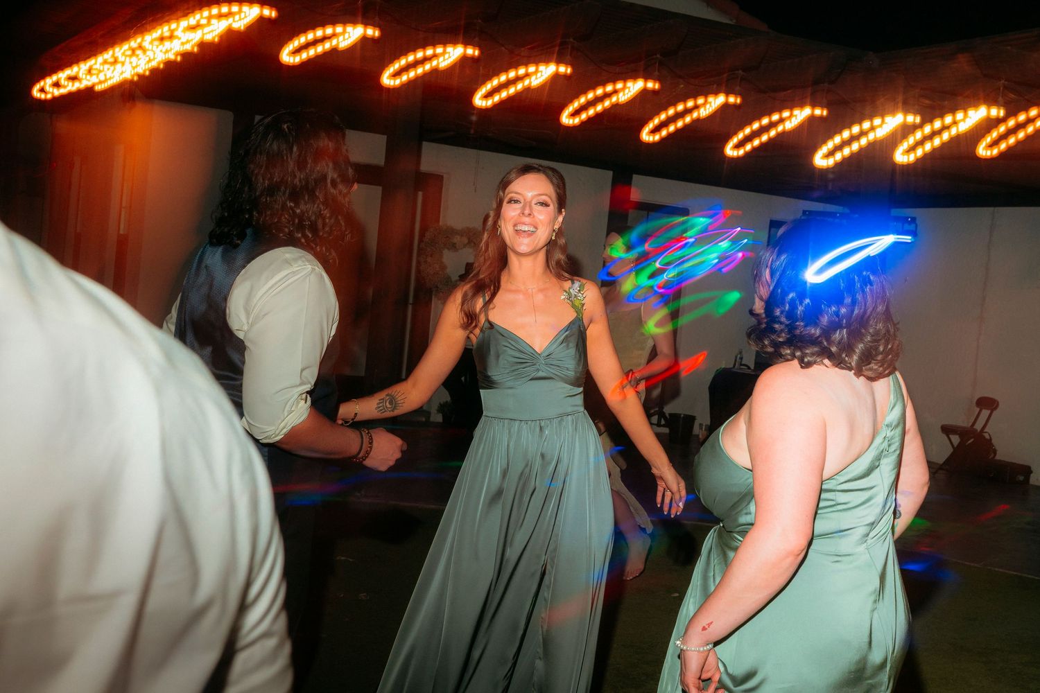Woman in sage green bridesmaid dress dancing at colorful wedding reception.
