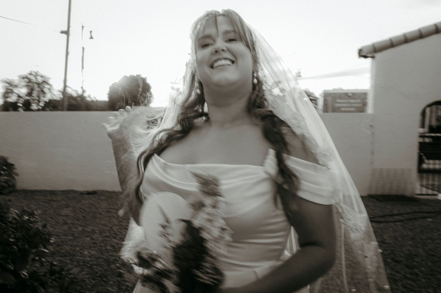 A smiling bride in a white wedding dress and veil holds a bouquet outdoors in this black and white photo.