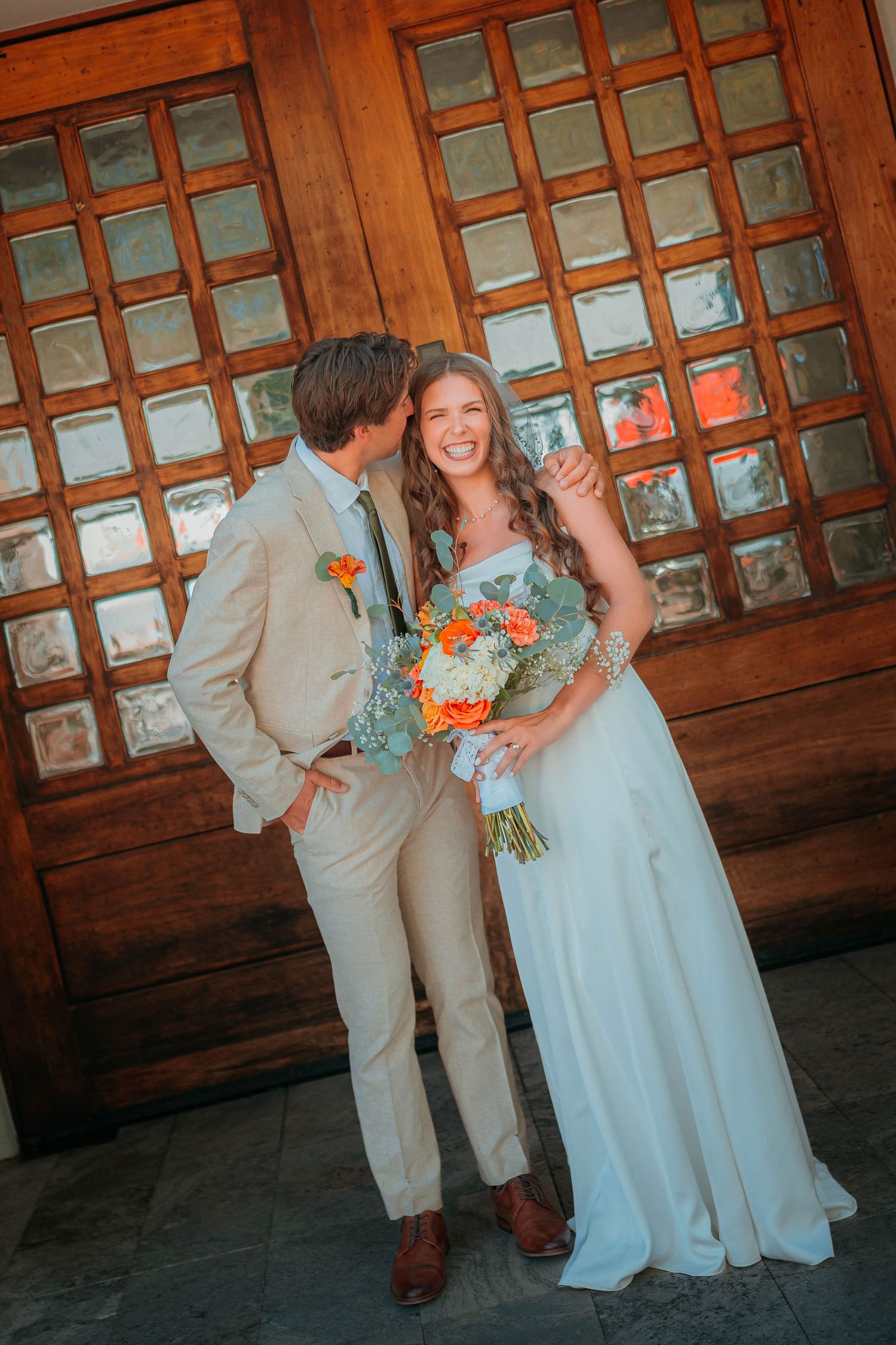 Bride in white gown holds orange bouquet as groom in tan suit kisses her cheek before wooden door.