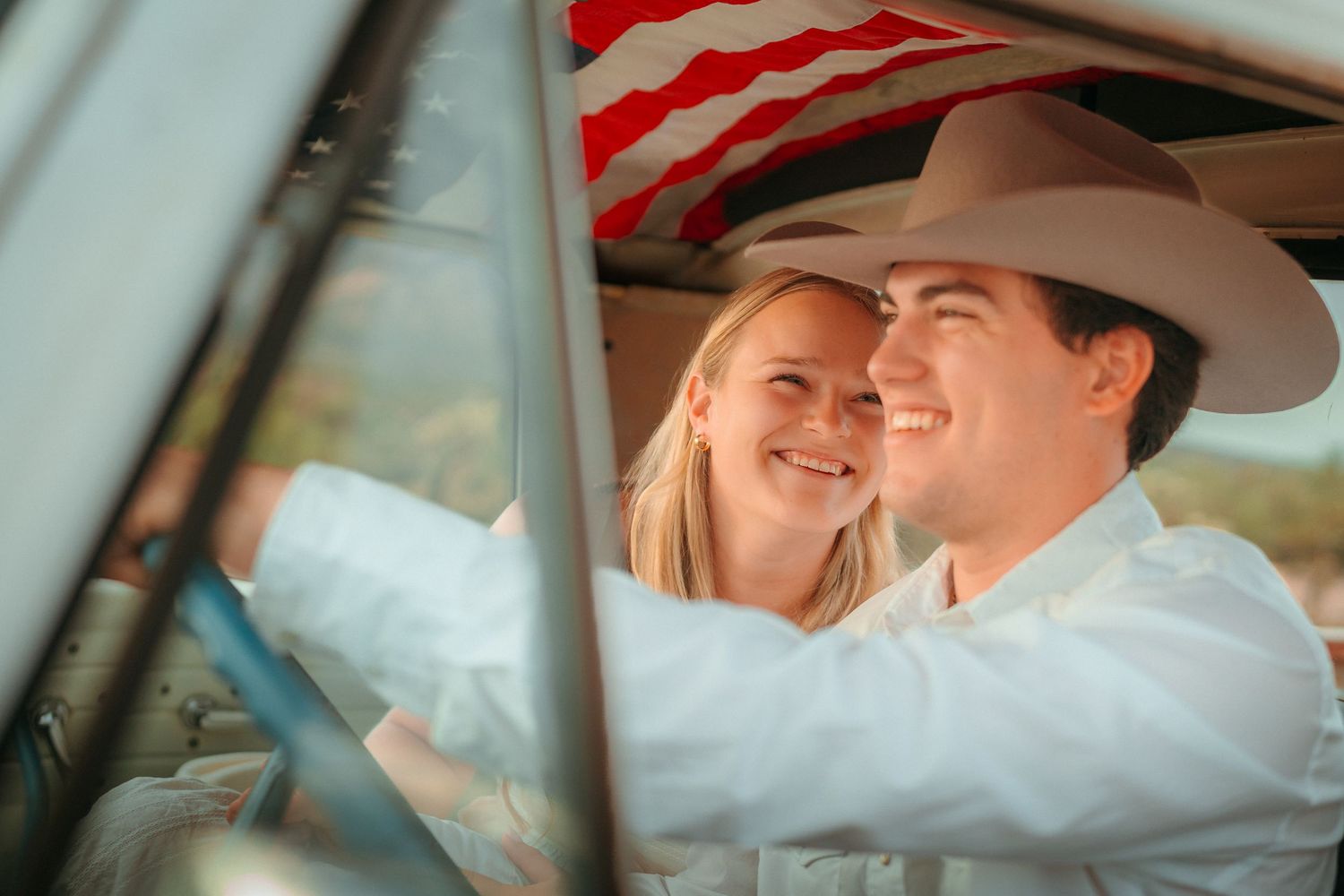 A smiling couple sits in a vintage truck cab with an American flag, man wearing cowboy hat and white shirt.