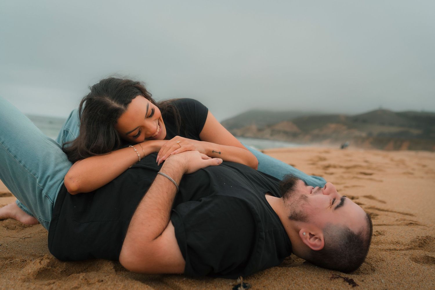 Playful couple lying on sandy beach, laughing together on a cloudy day by the ocean.