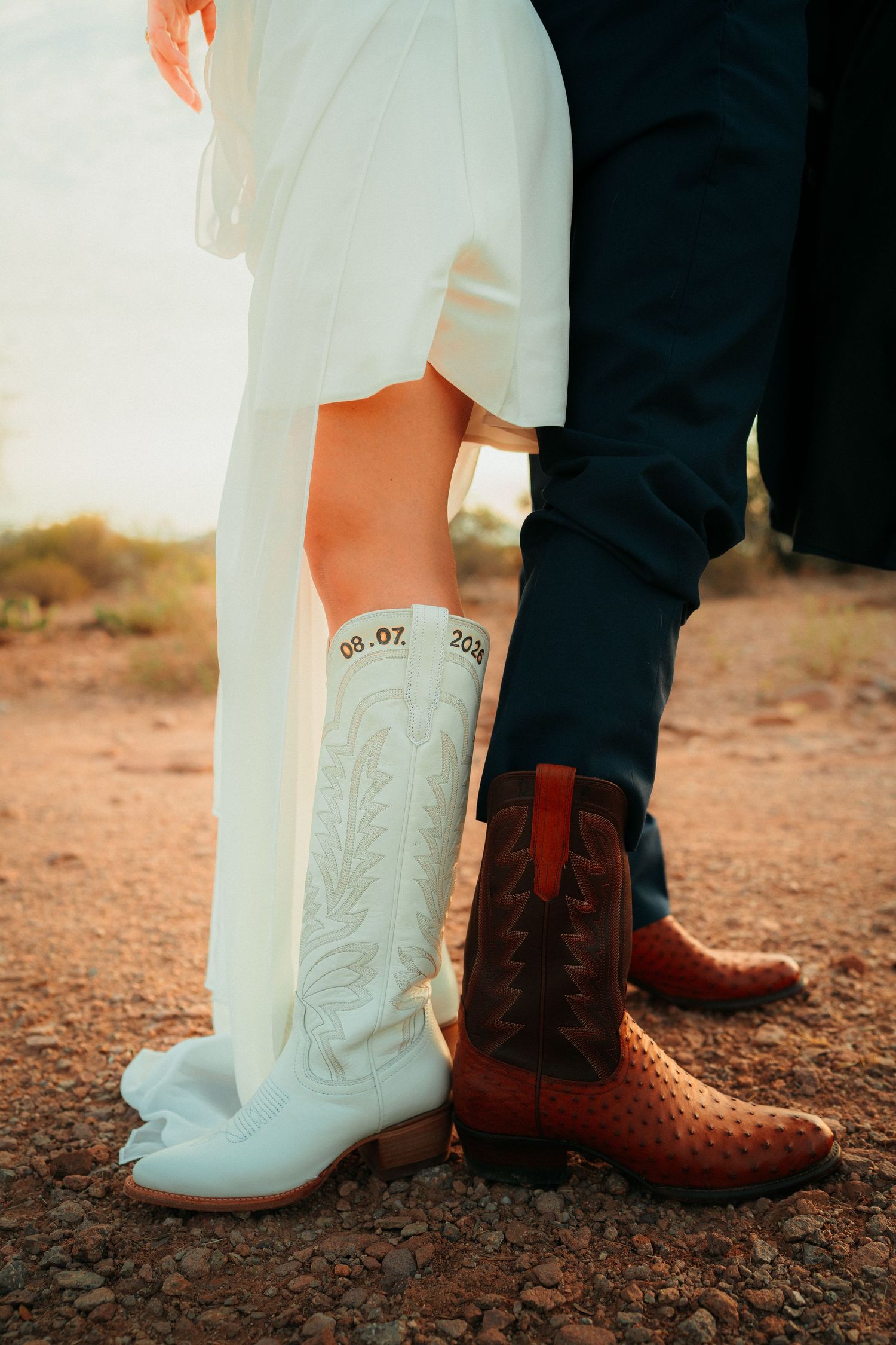 Bride and groom wearing custom cowboy boots with wedding date on red dirt ground, white bridal dress and navy suit.