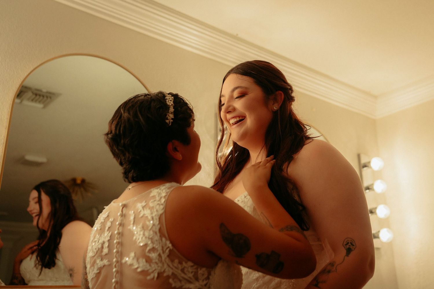 Two brides in white lace wedding dresses laughing together in a bridal suite.