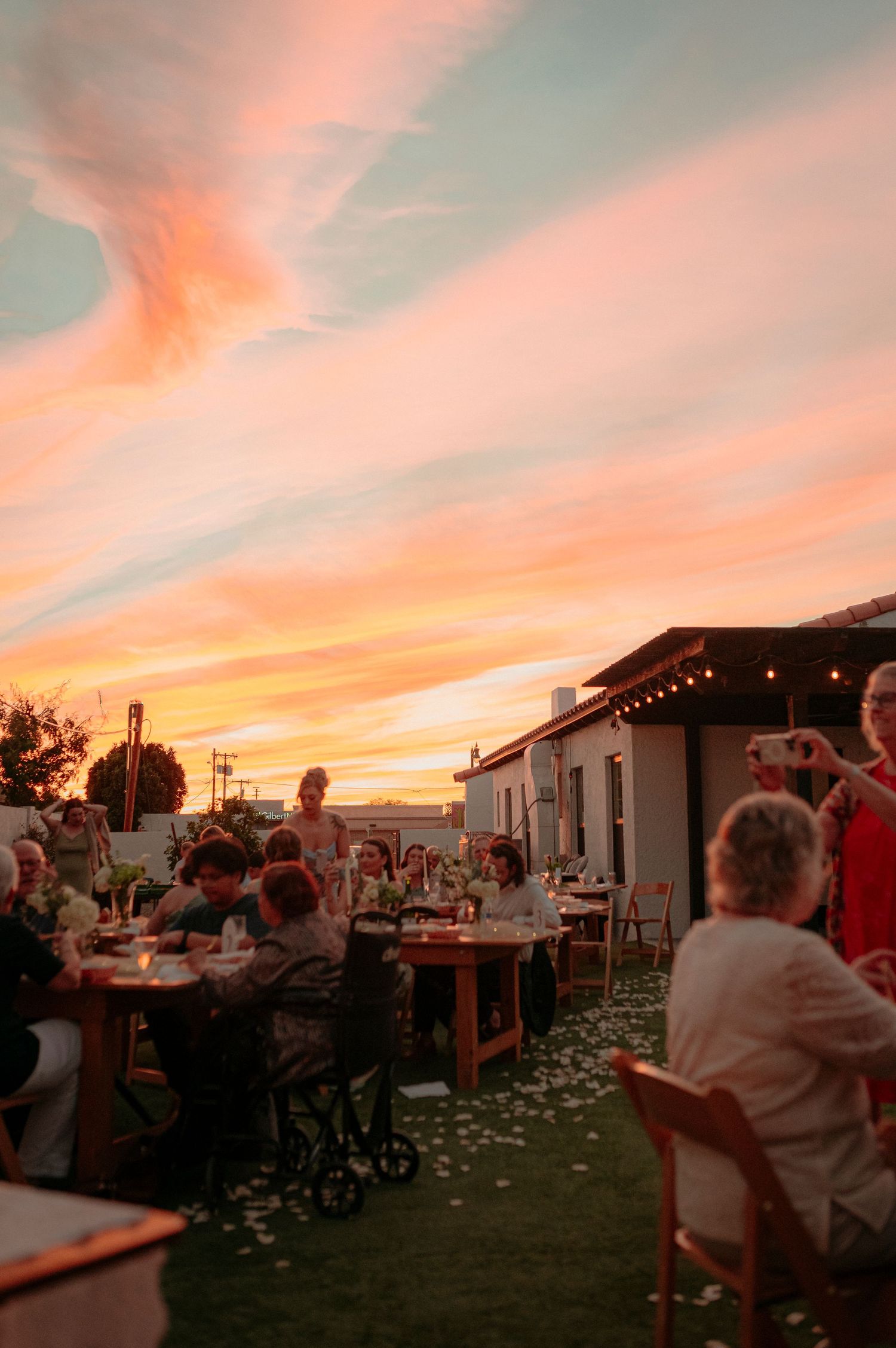Wedding guests dine at outdoor tables beneath a stunning pink and orange sunset sky.