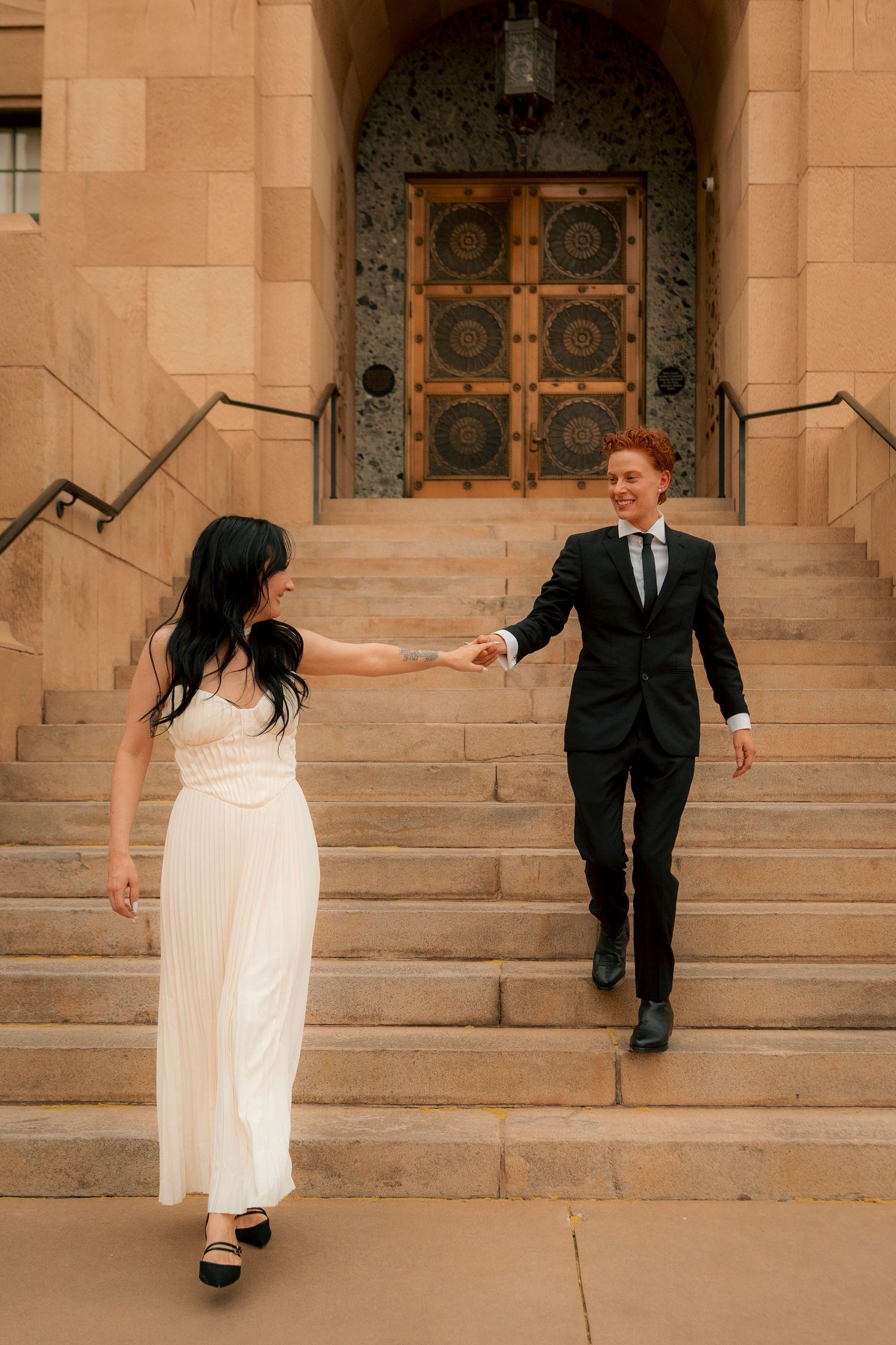 Bride in white dress and groom in black suit hold hands walking down stone steps outside a grand building.