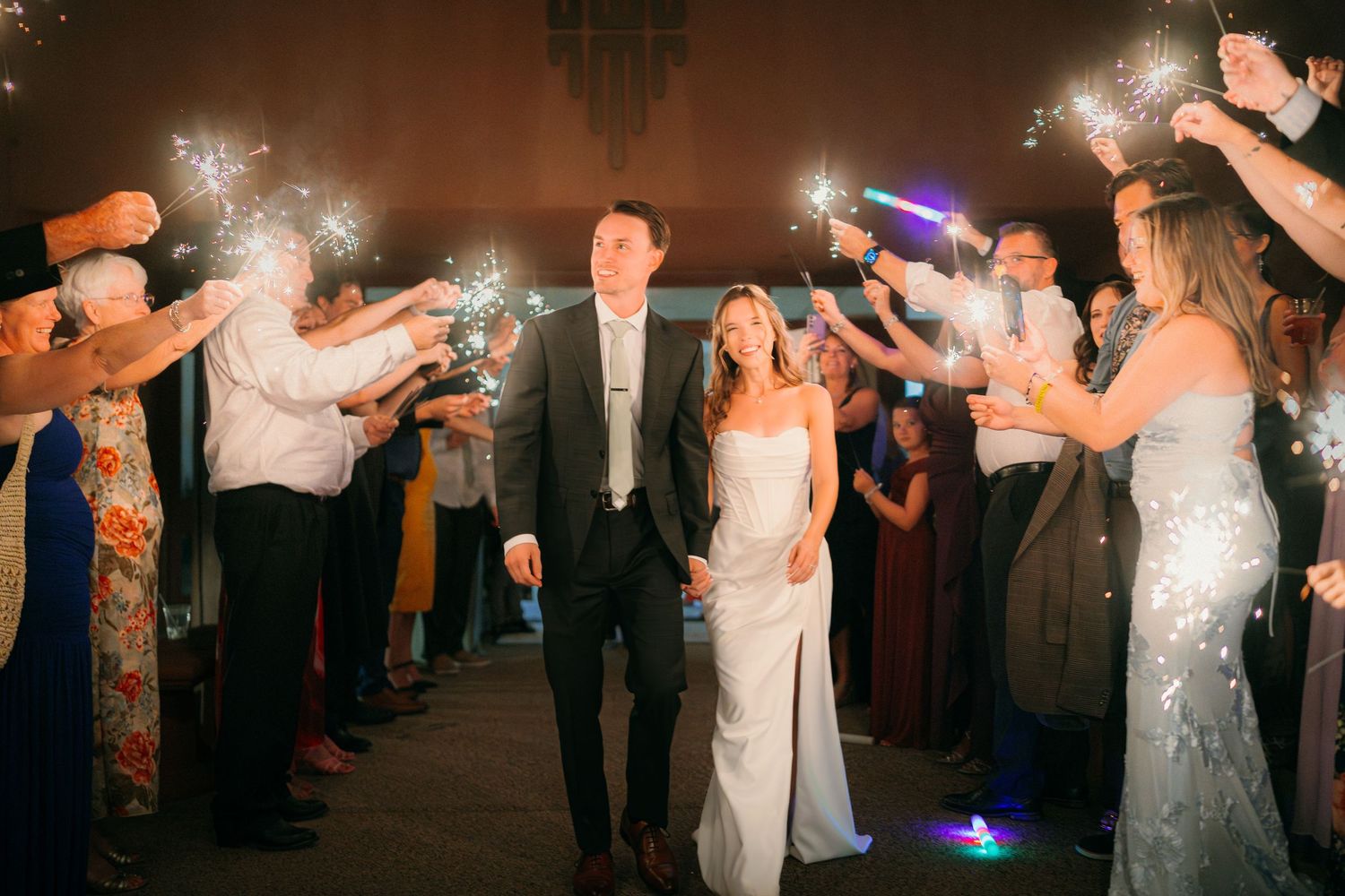Bride and groom exit wedding reception through sparkler tunnel surrounded by cheering guests at night.